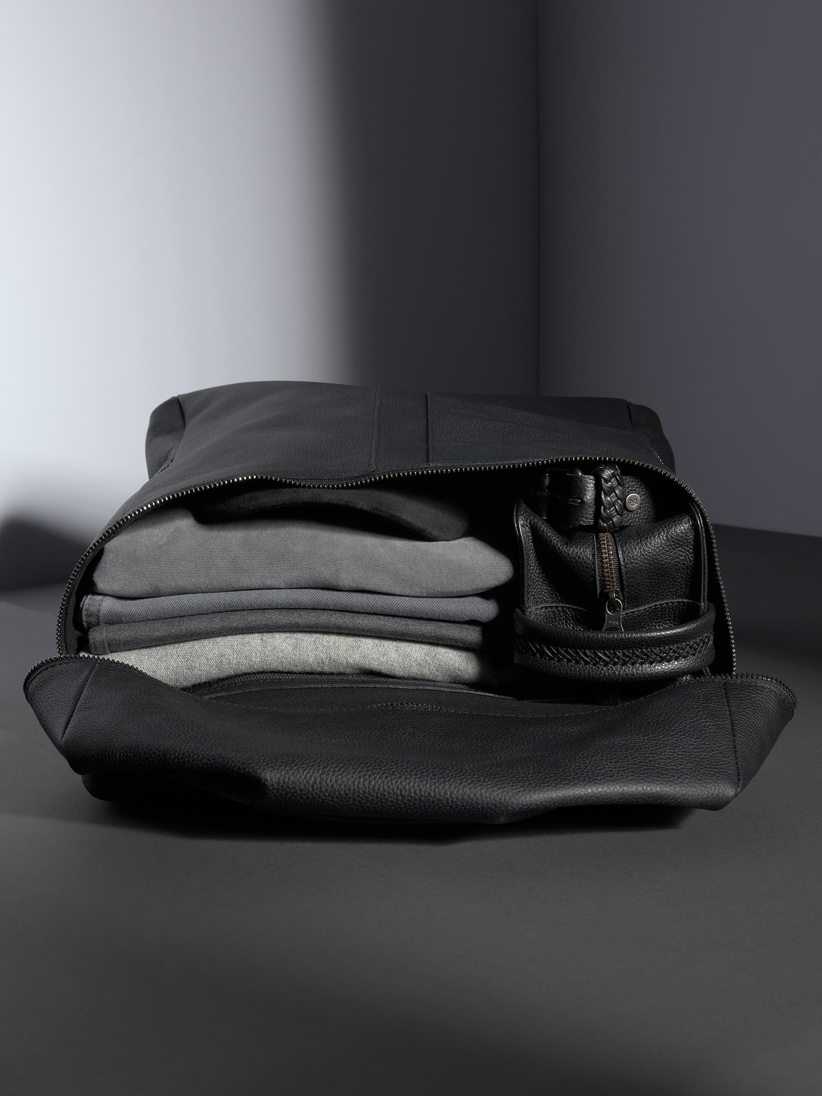Open Capra Leather Banteng black full-grain leather backpack showing neatly stacked gray clothing and a leather dopp kit.