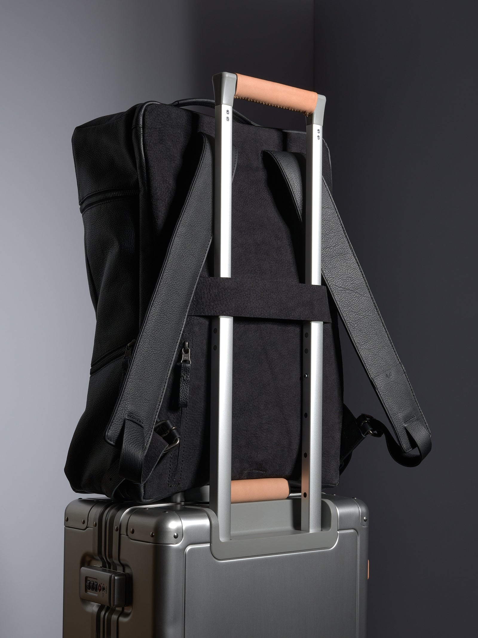Black leather backpack securely slid over the telescoping handle of a silver rolling suitcase for easy travel.