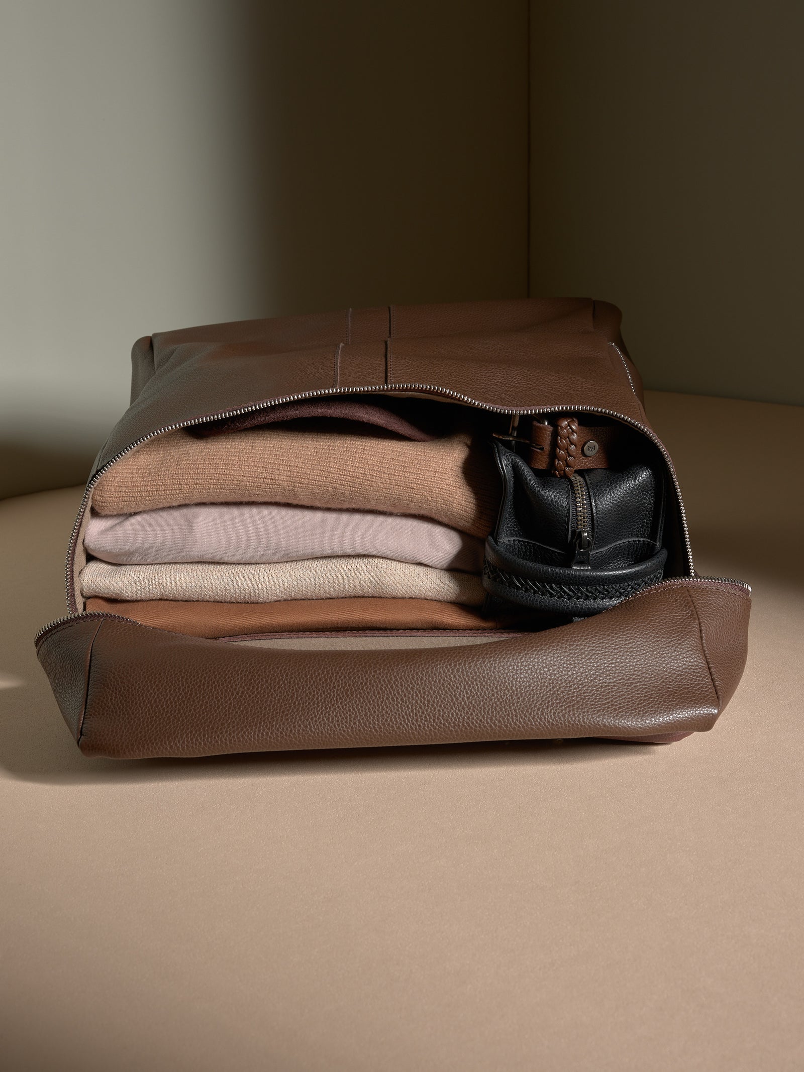 Open brown full-grain leather backpack showing organized travel essentials like folded clothes and a pouch.