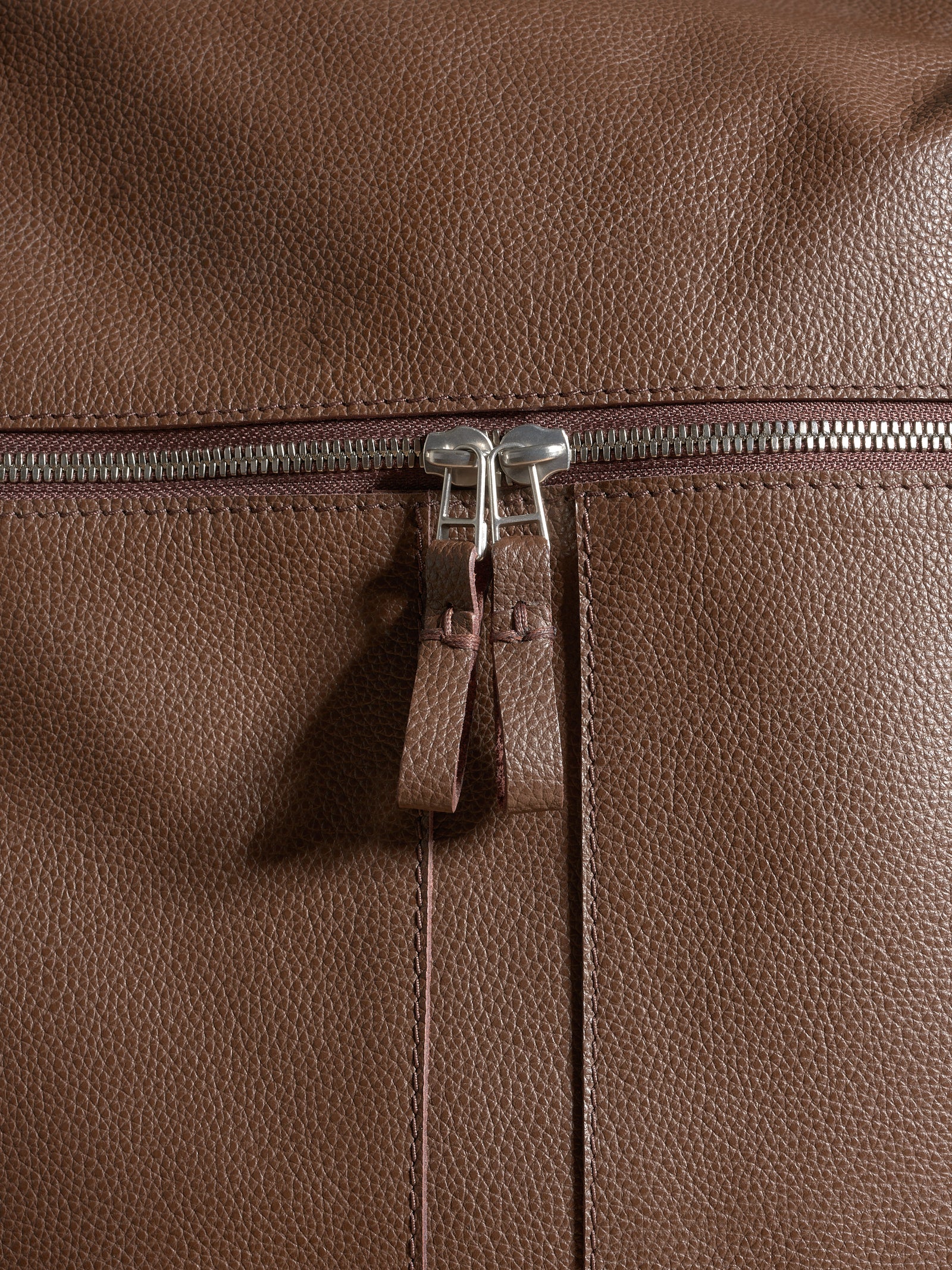 Extreme close-up of the textured brown full-grain leather, stitching, and durable metal zippers.