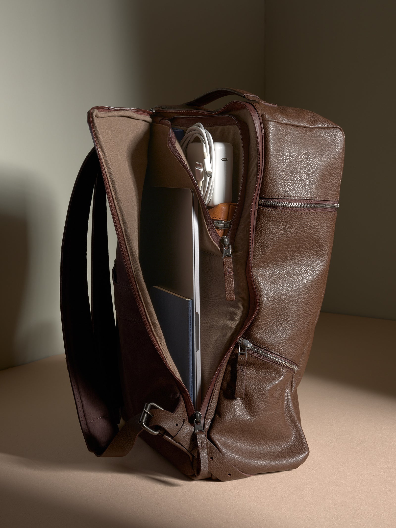 Open view of the brown leather backpack with a laptop, notebooks, and charging cables inside.