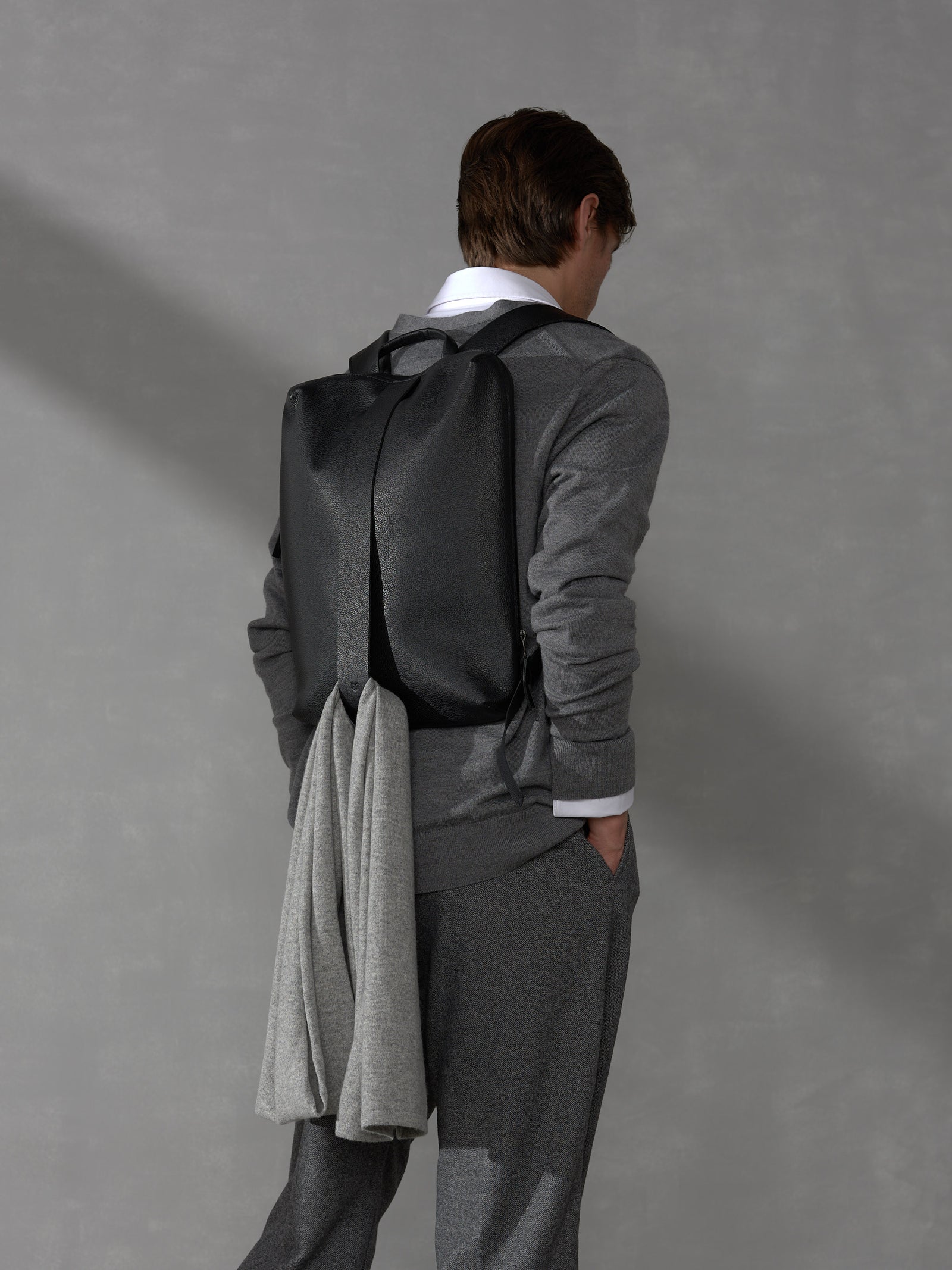Back view of a man wearing a black leather backpack with a grey knit sweater looped through the back strap.
