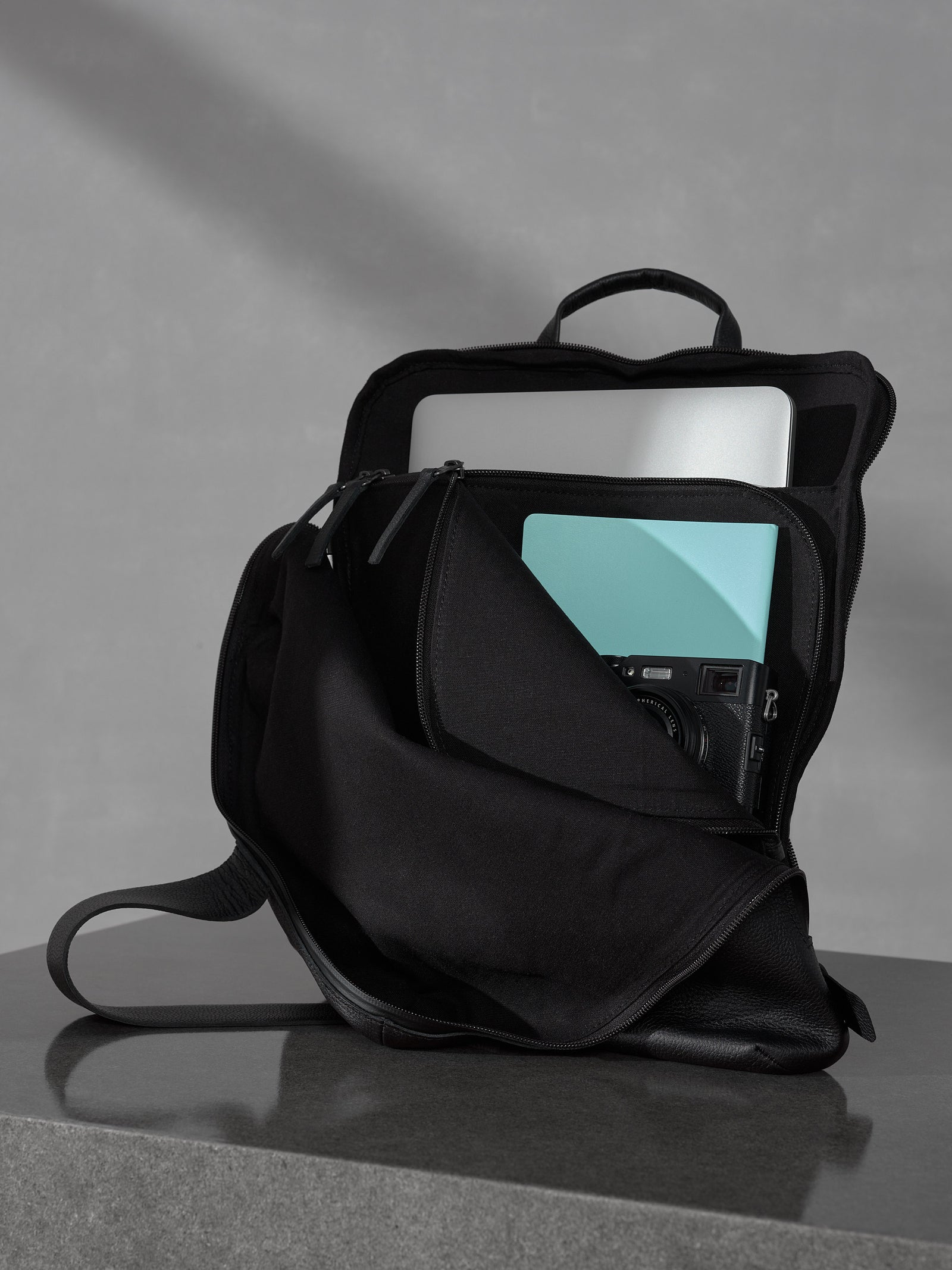 Open black leather backpack interior organizing a laptop, a teal notebook, and a professional mirrorless camera.