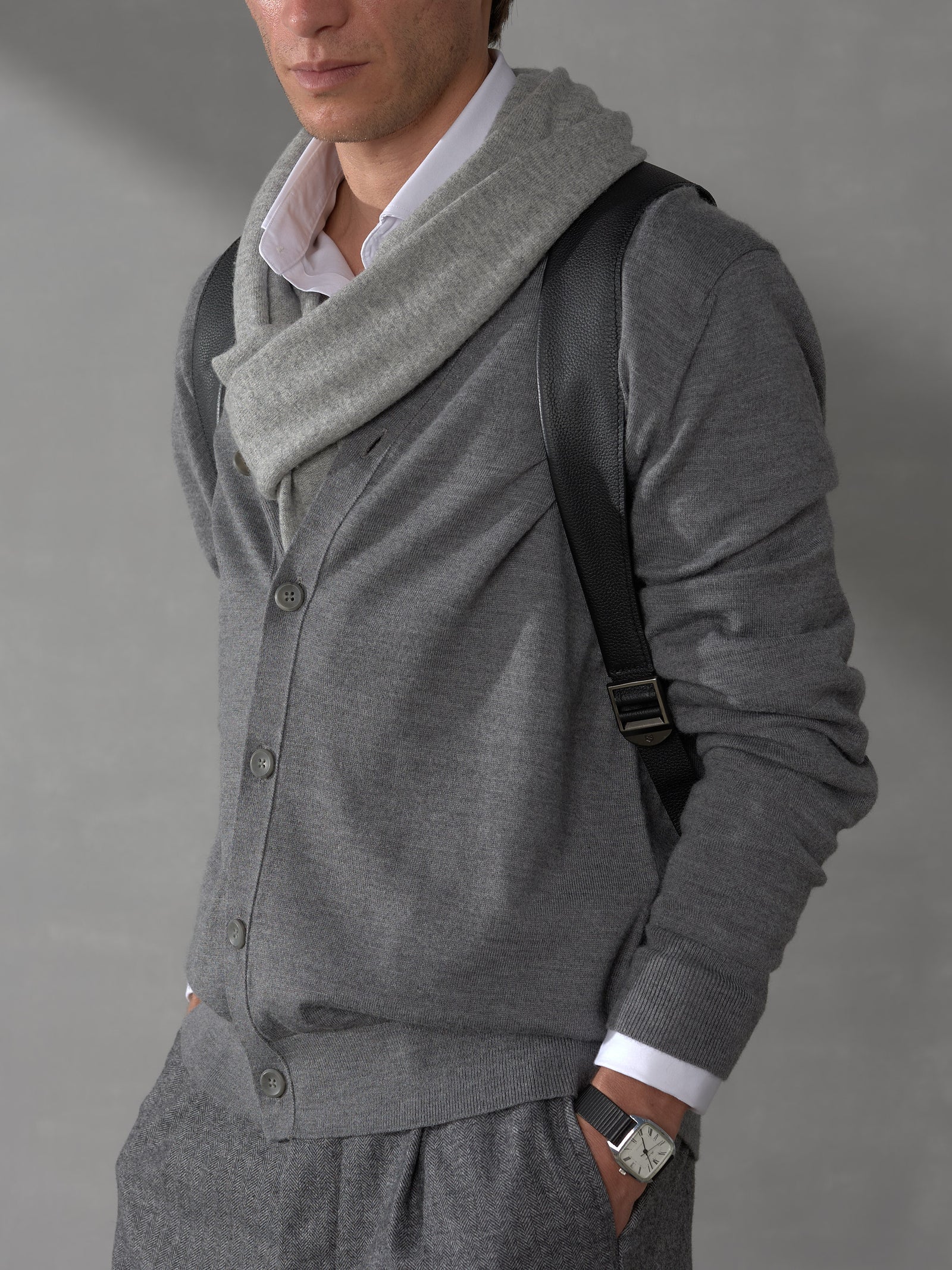 Close-up of a man in a grey cardigan wearing a black leather backpack strap with sleek metal hardware adjustment.