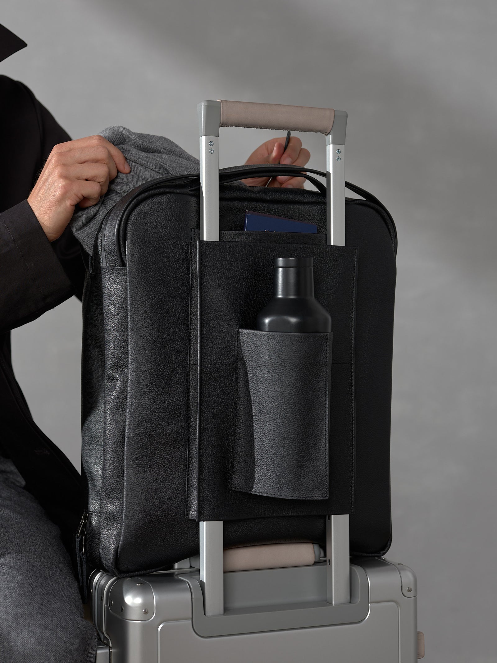 Close-up of a black leather backpack trolley sleeve secured on a suitcase handle, featuring a water bottle pocket.