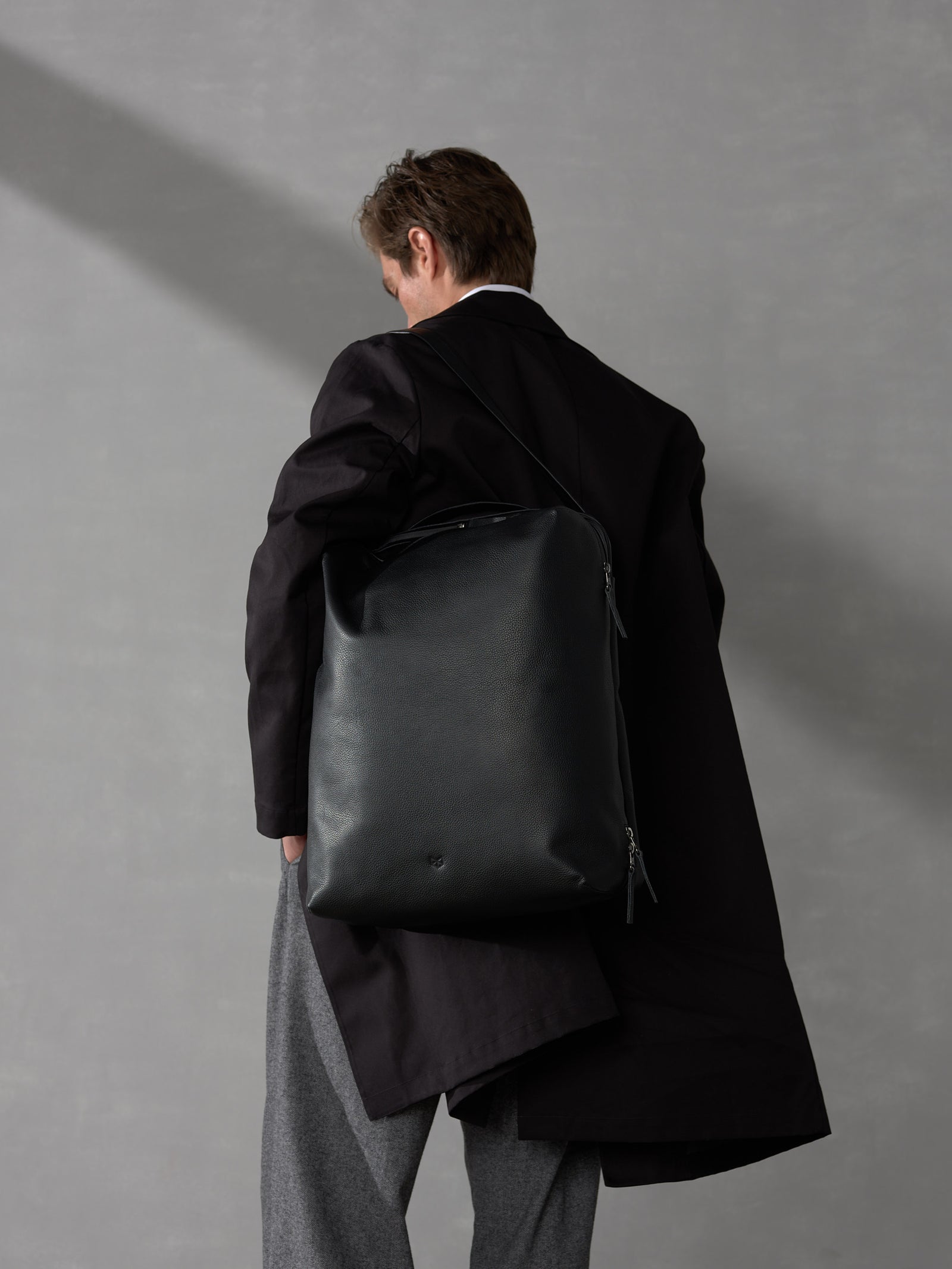 Back view of a man in a dark coat carrying a sleek, minimalist black leather backpack on one shoulder.