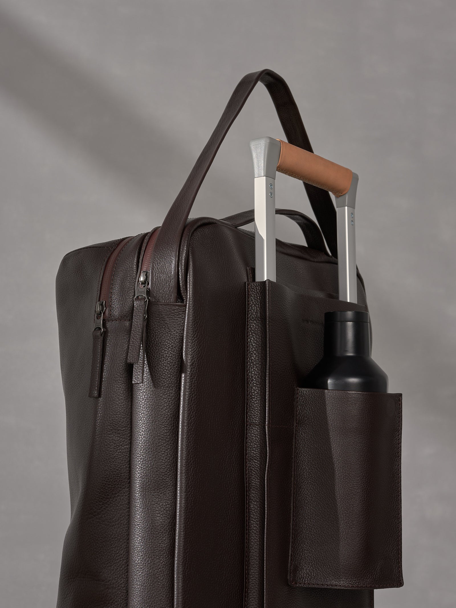 Close-up of a Capra Leather backpack strapped to a luggage handle featuring a built-in water bottle pocket.