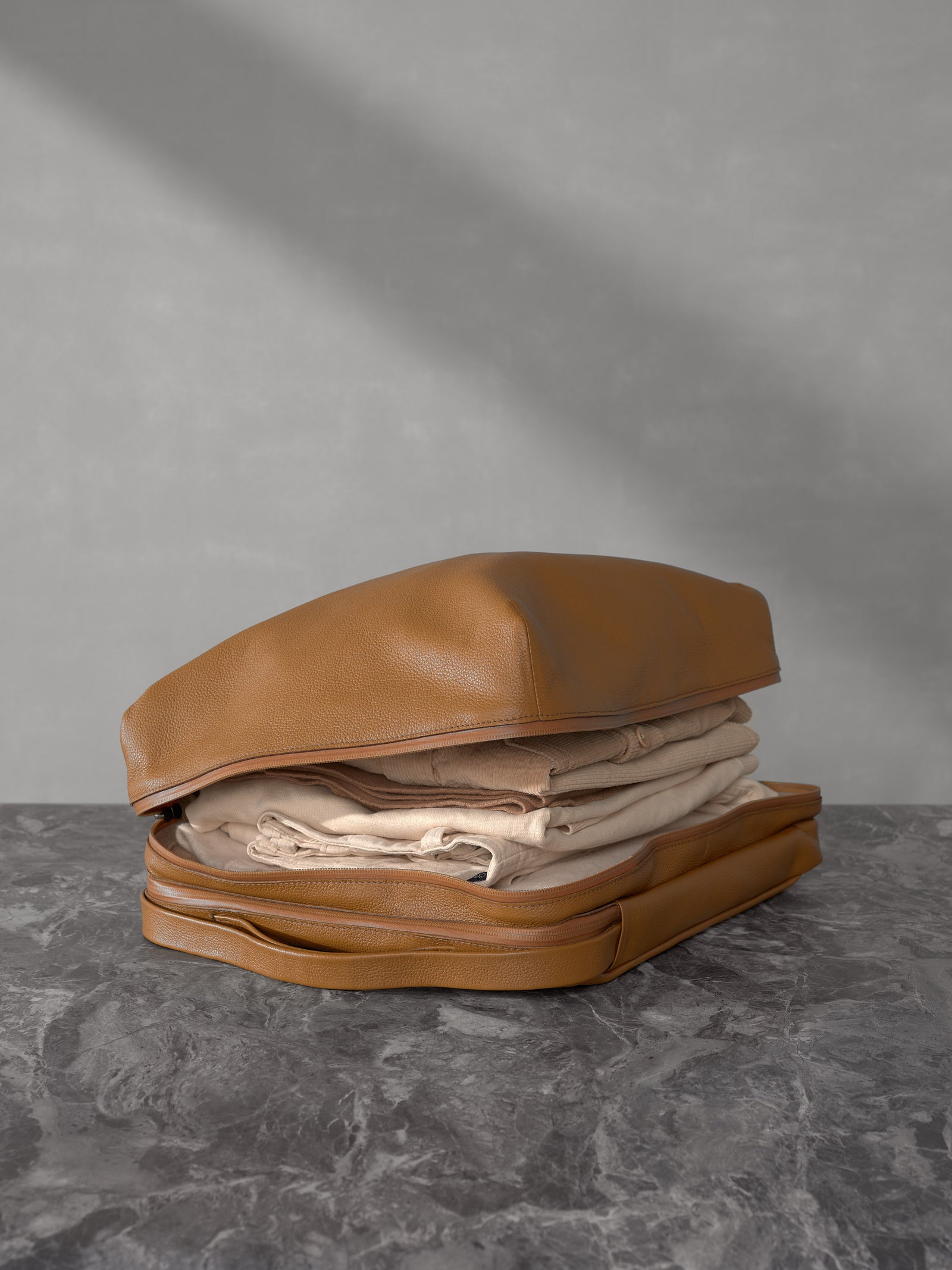 Open tan leather travel organizer pouch from Capra Leather packed with folded cream-colored clothing on a dark marble surface.