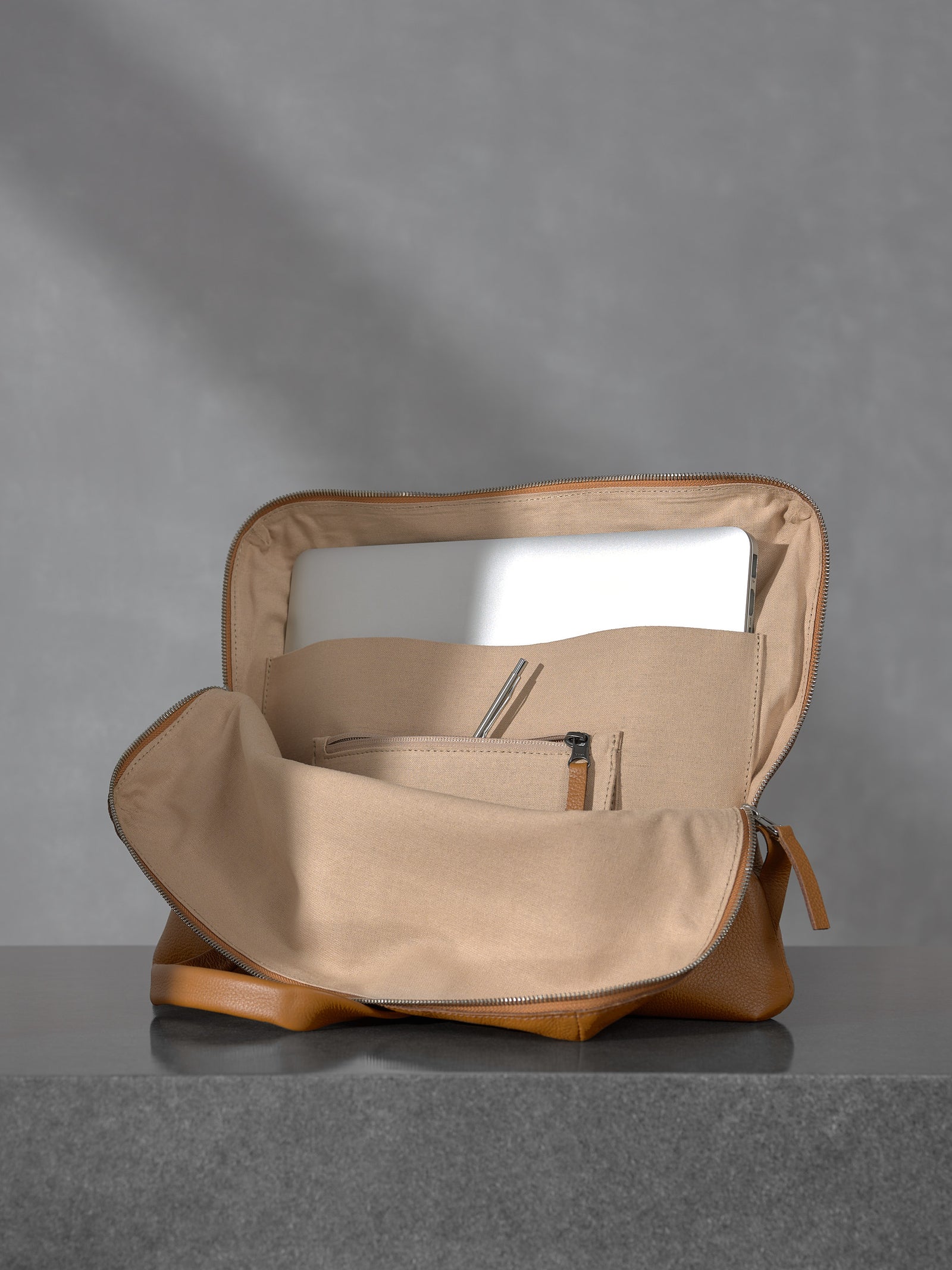 Internal view of the open Capra Leather tan briefcase showing a dedicated laptop compartment holding a silver MacBook, canvas lining, and zippered interior pockets.
