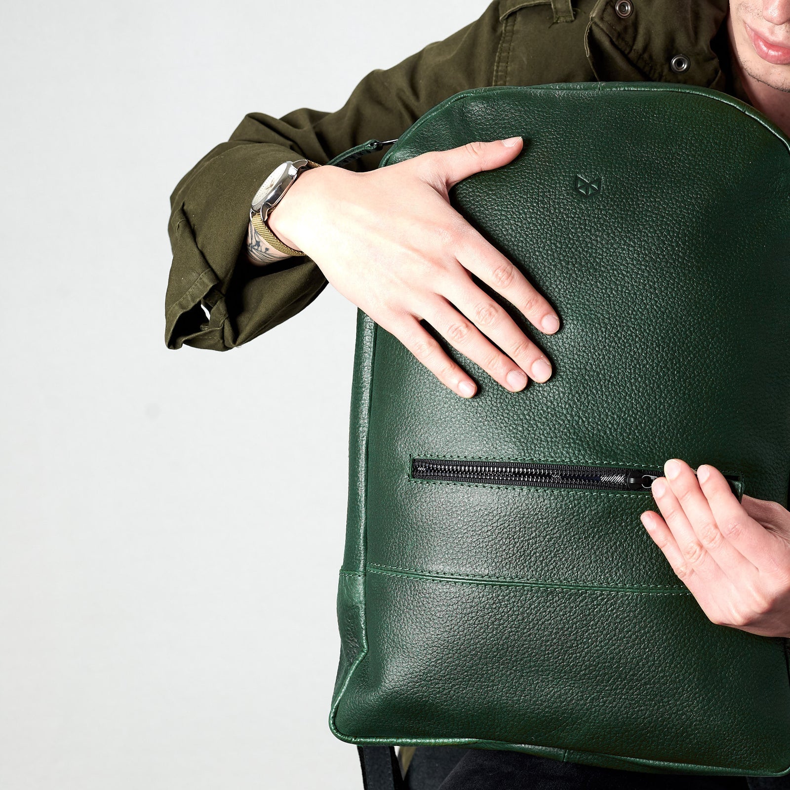 A close-up shot of a person's hands embracing the front of the green, pebble-grain leather backpack, highlighting the textured material and the horizontal zipper pocket.