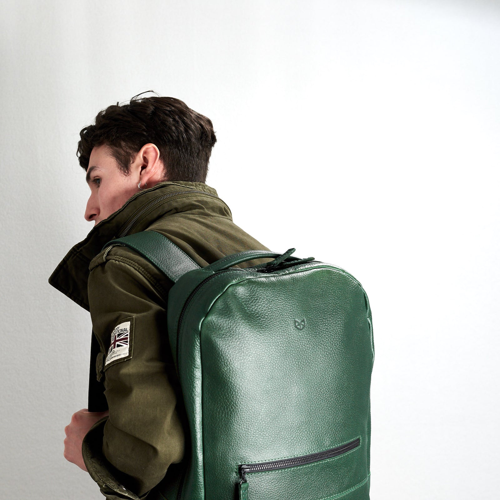 A side view of a young man wearing an olive green jacket with the green, pebble-grain leather backpack on his back, emphasizing the rounded shape and texture.