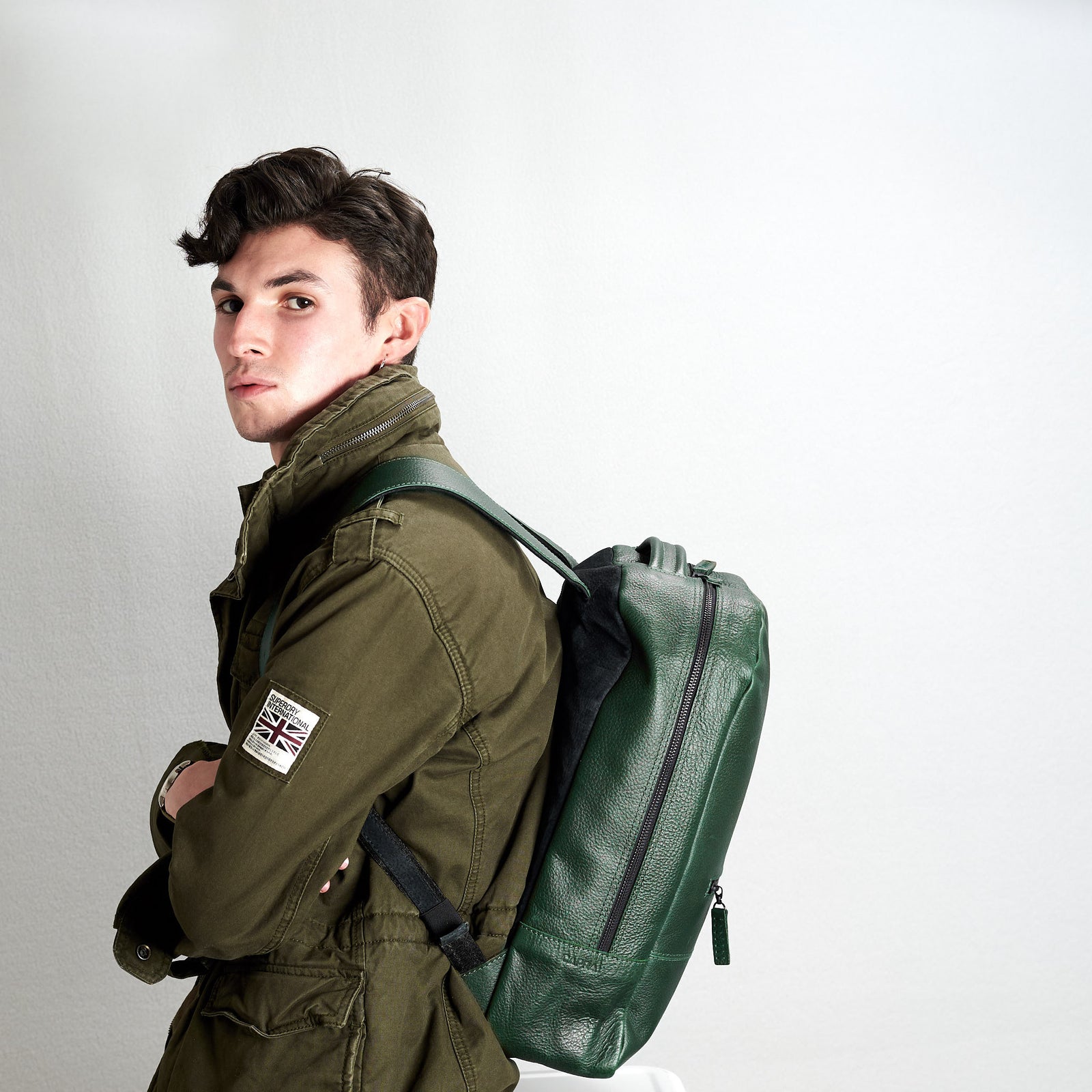 A young man wearing an olive green jacket looks over his shoulder while carrying a deep green, pebble-grain leather backpack.