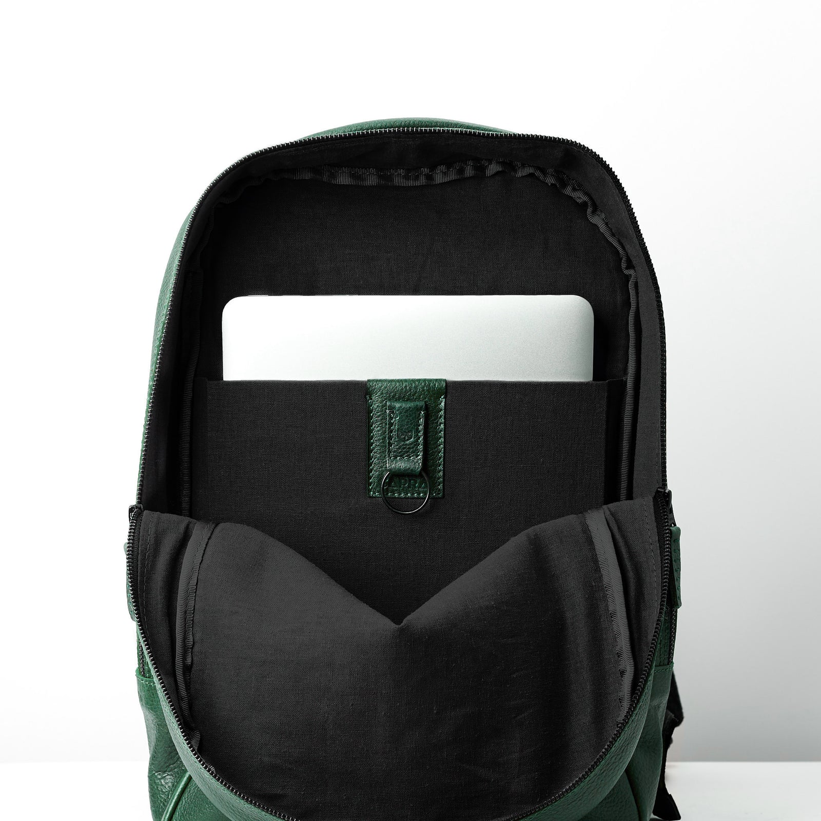 The interior of an open green leather backpack. A silver laptop is visible in the black padded sleeve, which is secured by a green leather tab.