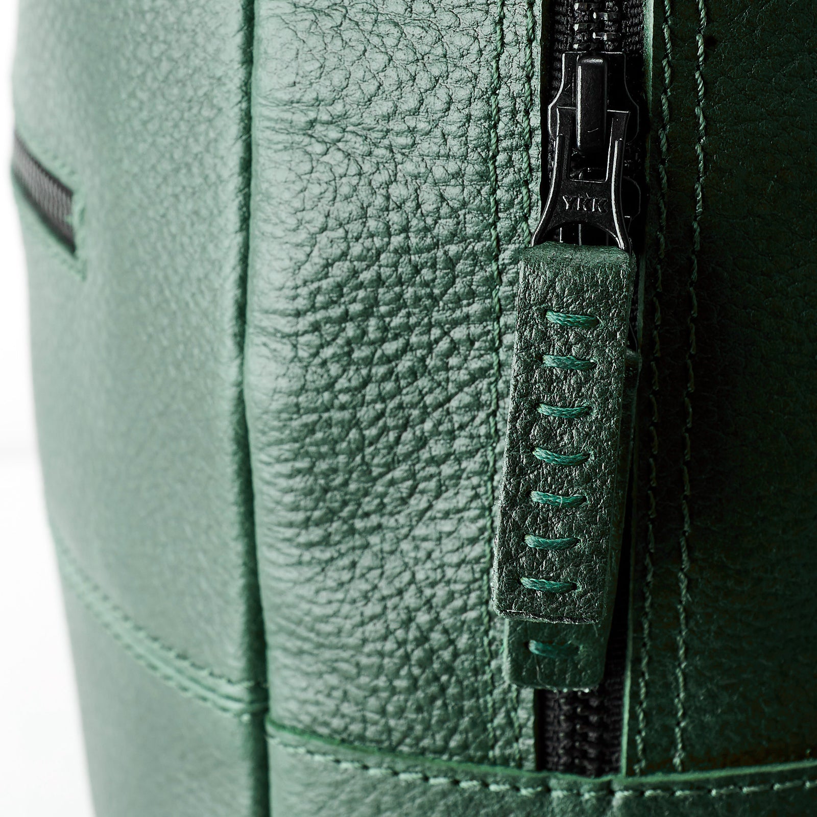 An extreme close-up detail of the black YKK zipper and the green leather zipper pull tab with visible dark green stitching on the pebble-grain leather backpack.