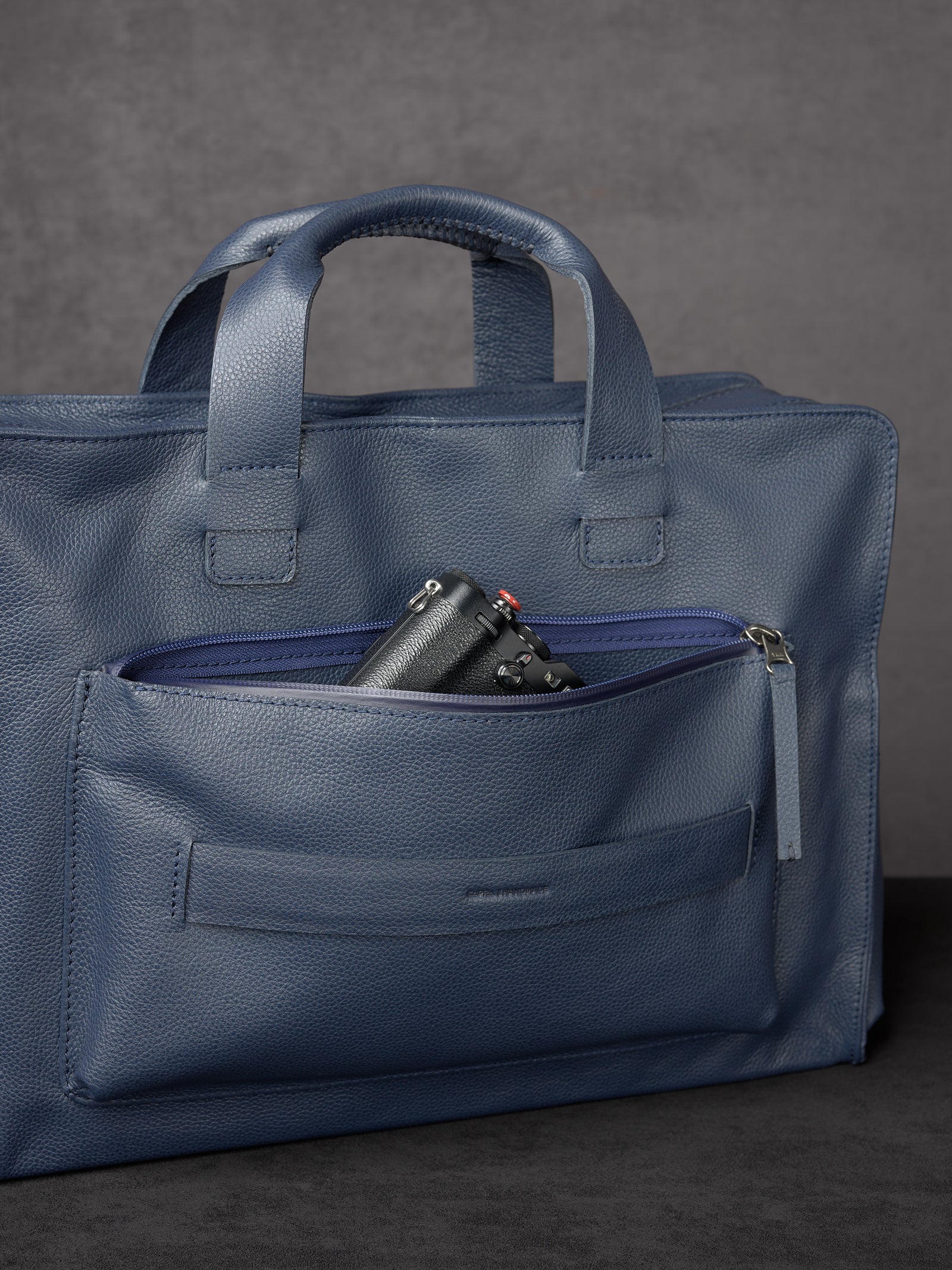 A black compact camera tucked into the zippered front pouch of a navy blue leather briefcase.