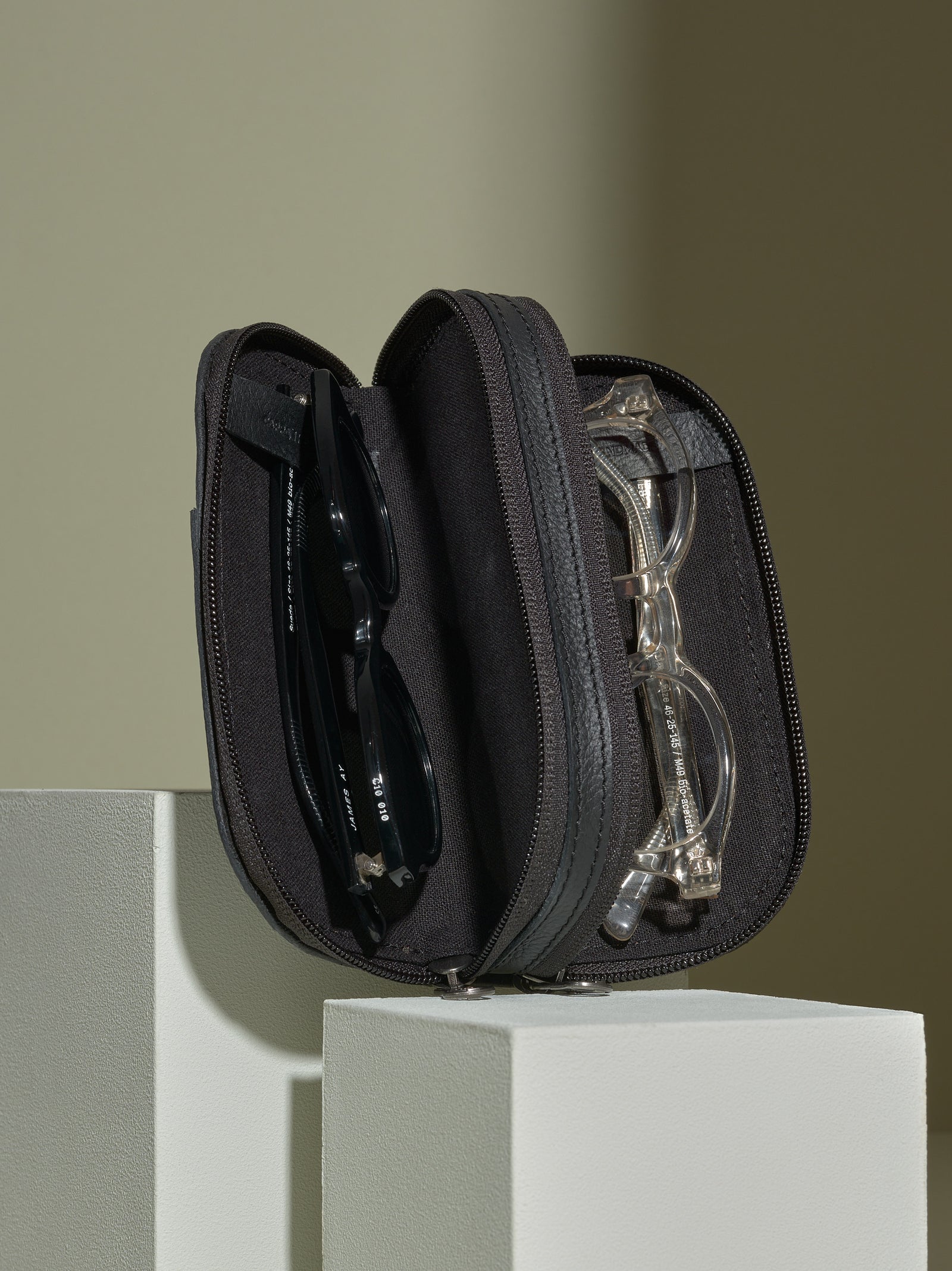 Black leather double-glasses case opened on white blocks, showing a pair of sunglasses and reading glasses inside.