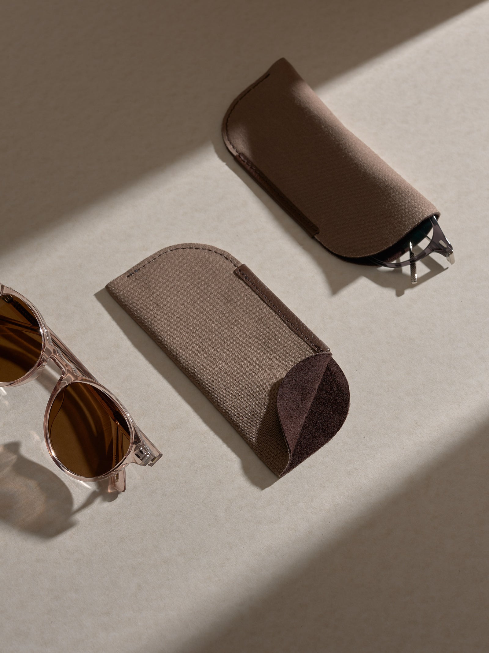 Two separate soft brown fabric sleeves, designed to hold glasses, are shown on a light-colored surface with shadows, next to a pair of clear pink-framed sunglasses.