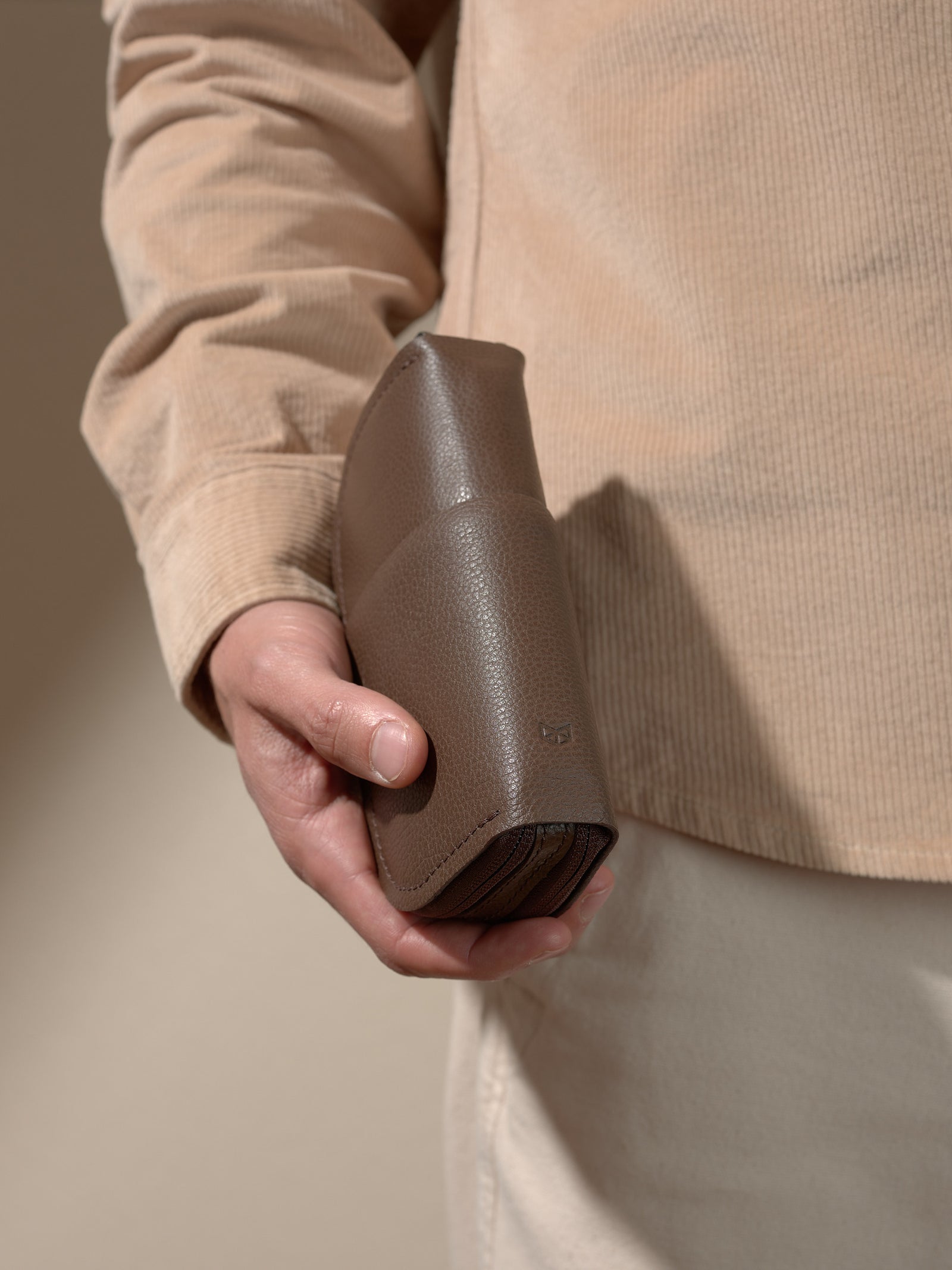 A person's hand holding the textured brown leather double glasses case, emphasizing the curved shape and pebbled leather texture.