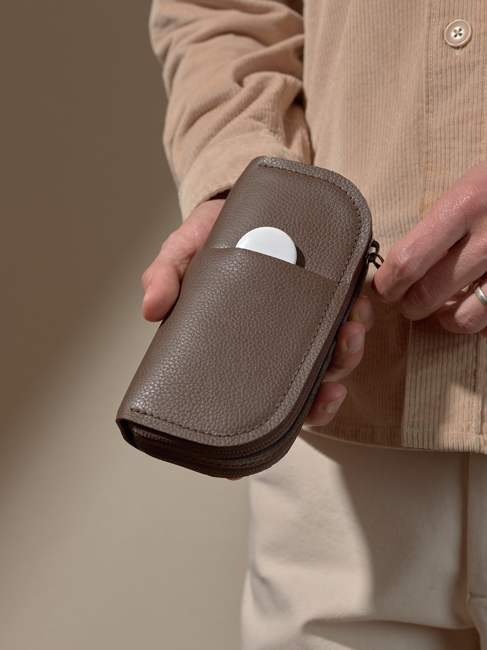 A person's hands holding the closed, textured brown leather glasses case and placing a white, circular object (like a tracker or earbud case) into a discreet slip pocket on the front.