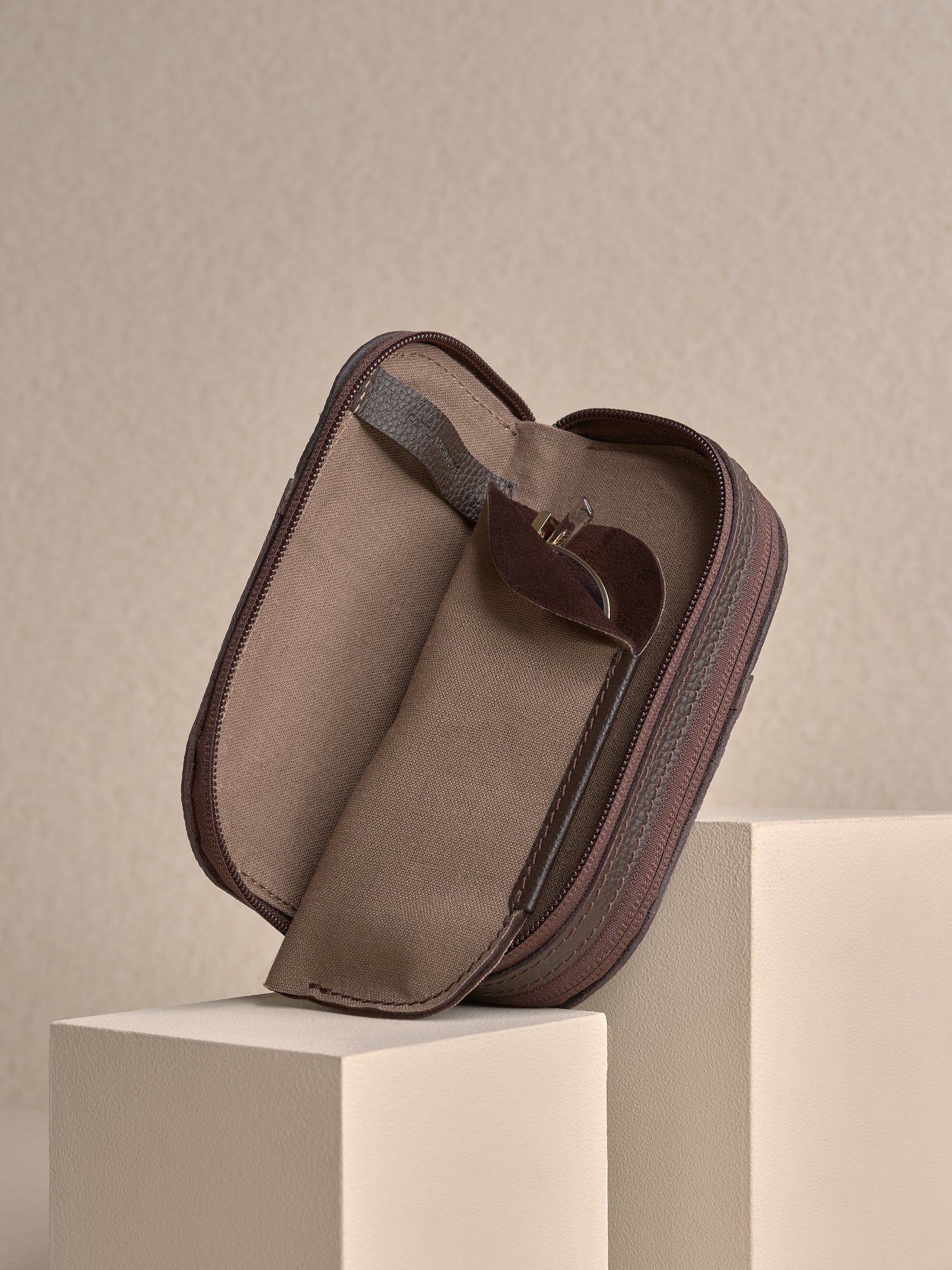 The opened, zipped dark brown leather glasses case standing upright on a white platform, showing one soft fabric sleeve inside with a pair of glasses partially inserted.