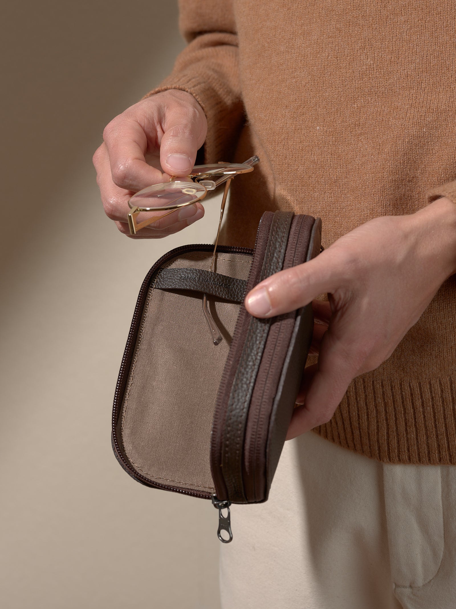 Close-up of a person's hands wearing a brown sweater, holding the opened dark brown leather case and removing or inserting a pair of gold-framed glasses.