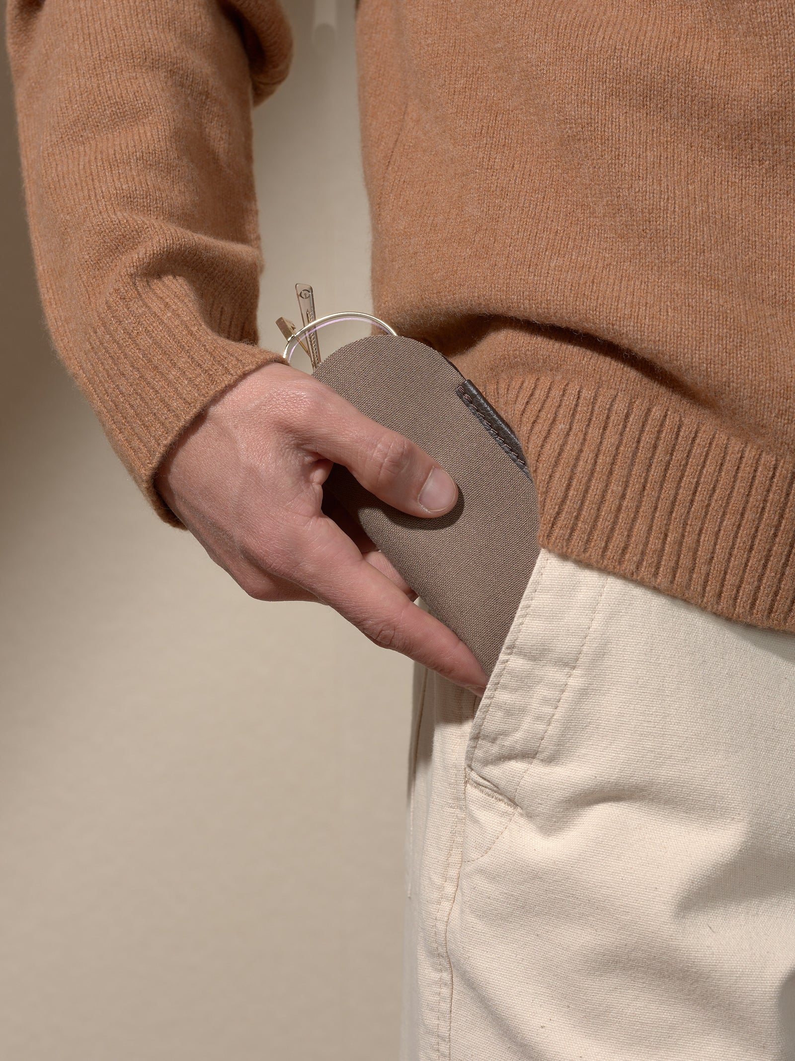Close-up of a person's hand inserting the soft dark brown fabric inner sleeve of the glasses case into the pocket of light-colored trousers, while wearing a brown sweater.