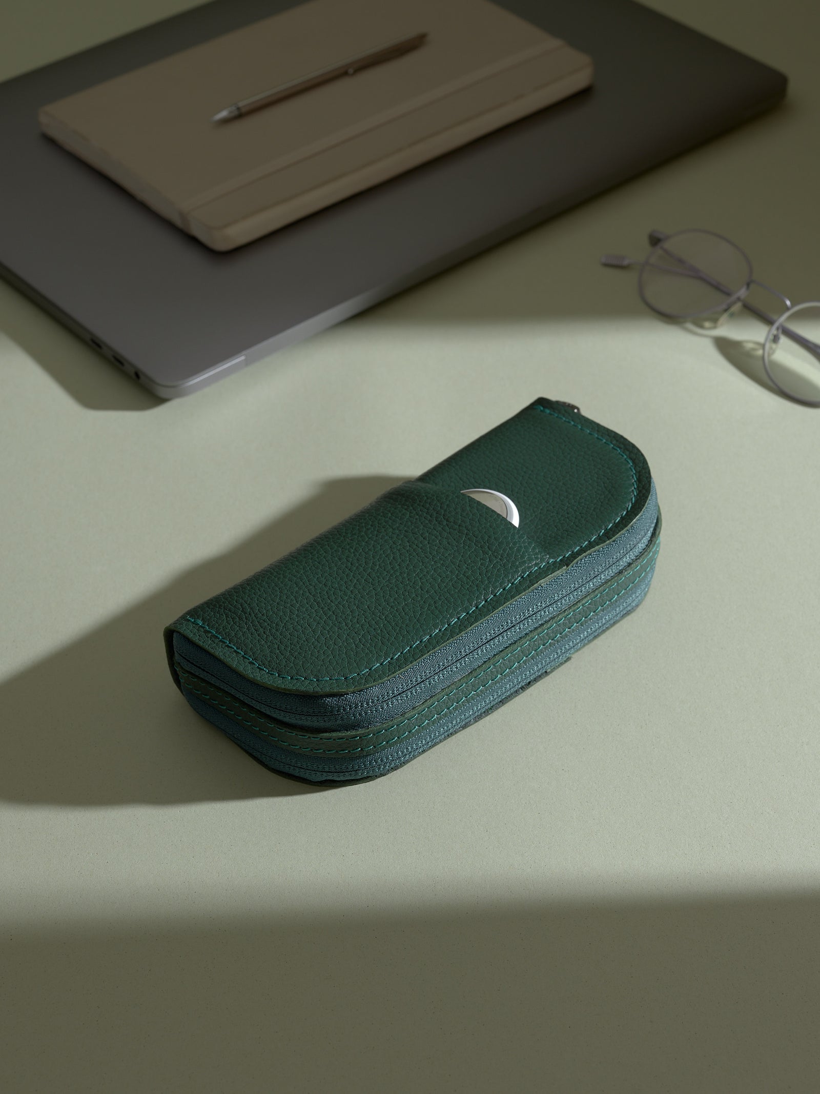 The closed, green Capra leather double glasses case resting on a light green desk, with a white circular object (likely an AirTag or earbud case) partially visible in the exterior slip pocket. A laptop, notepad, and glasses are in the background.