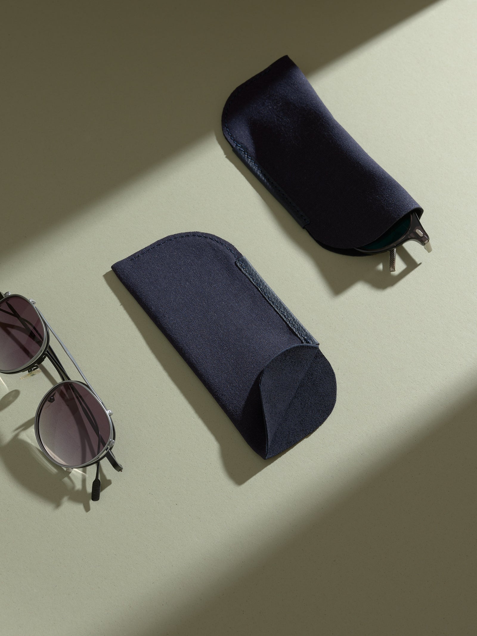 Two separate dark navy blue fabric sleeves, designed to hold glasses, are displayed on a light green surface next to a pair of round, silver-framed sunglasses.