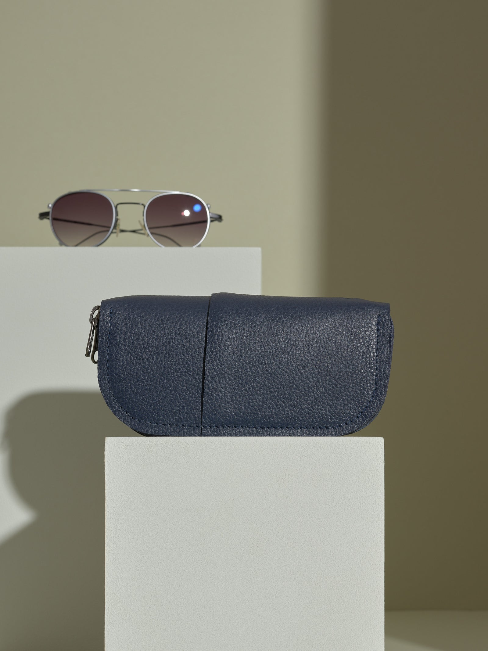 The closed, navy blue Capra leather double glasses case resting horizontally on two white platforms, with a pair of silver-framed aviator sunglasses positioned behind it on the upper platform.