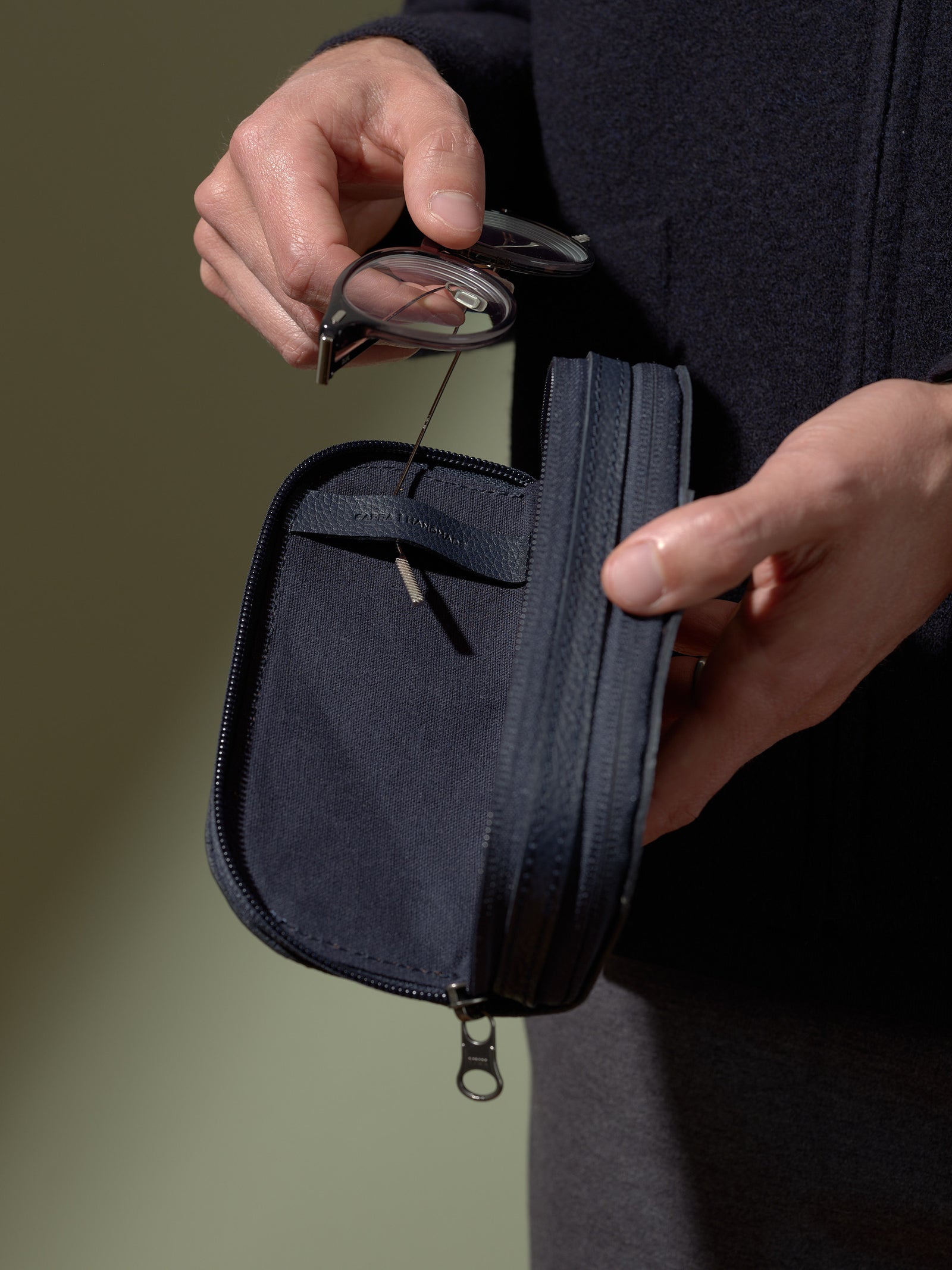 A person's hands holding the opened, navy blue leather glasses case and removing a pair of clear glasses from the soft inner compartment.