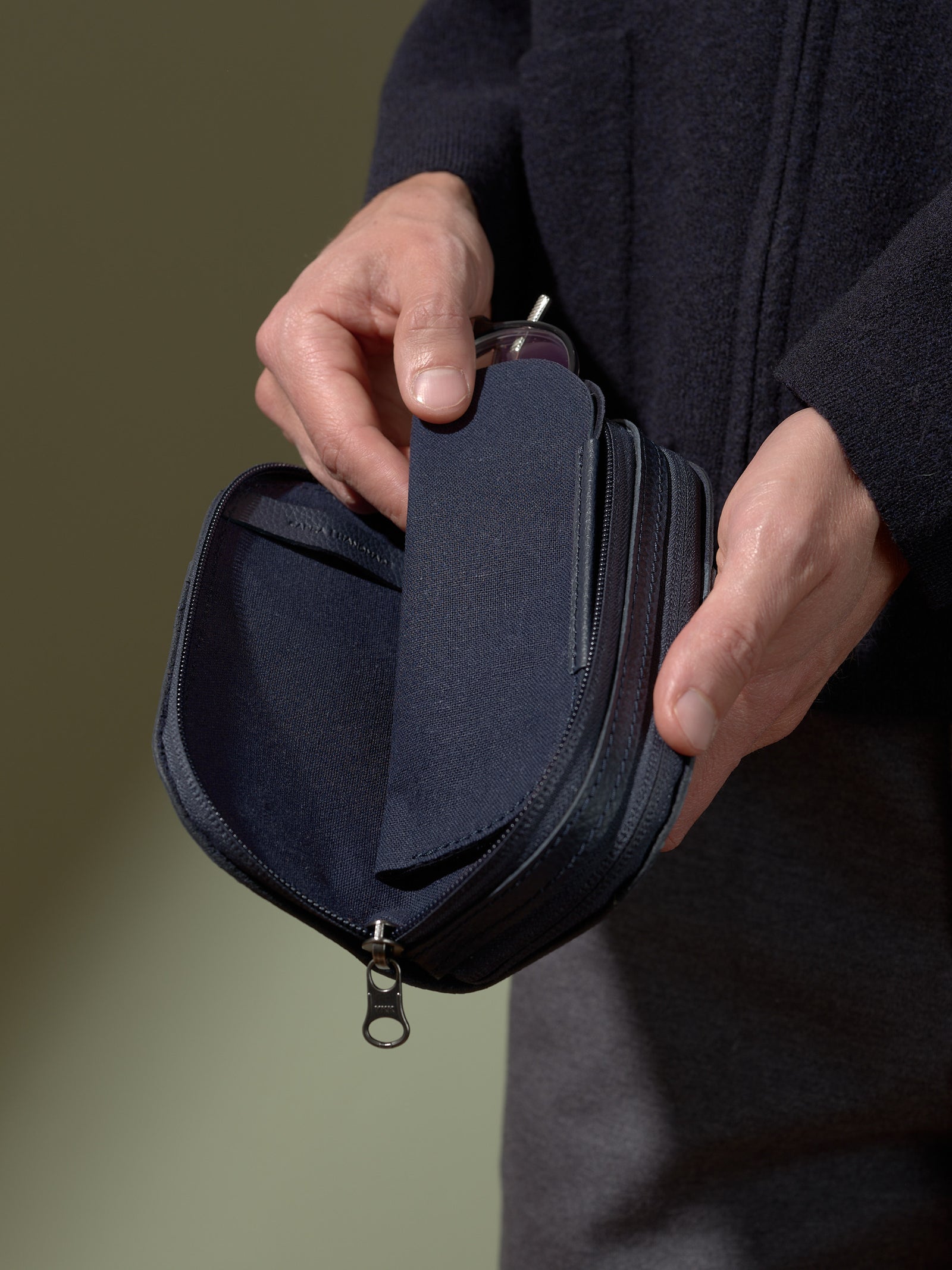 A close-up of a person's hands wearing a dark sweater, holding the opened navy blue leather case and removing a pair of glasses contained in the soft fabric inner sleeve.