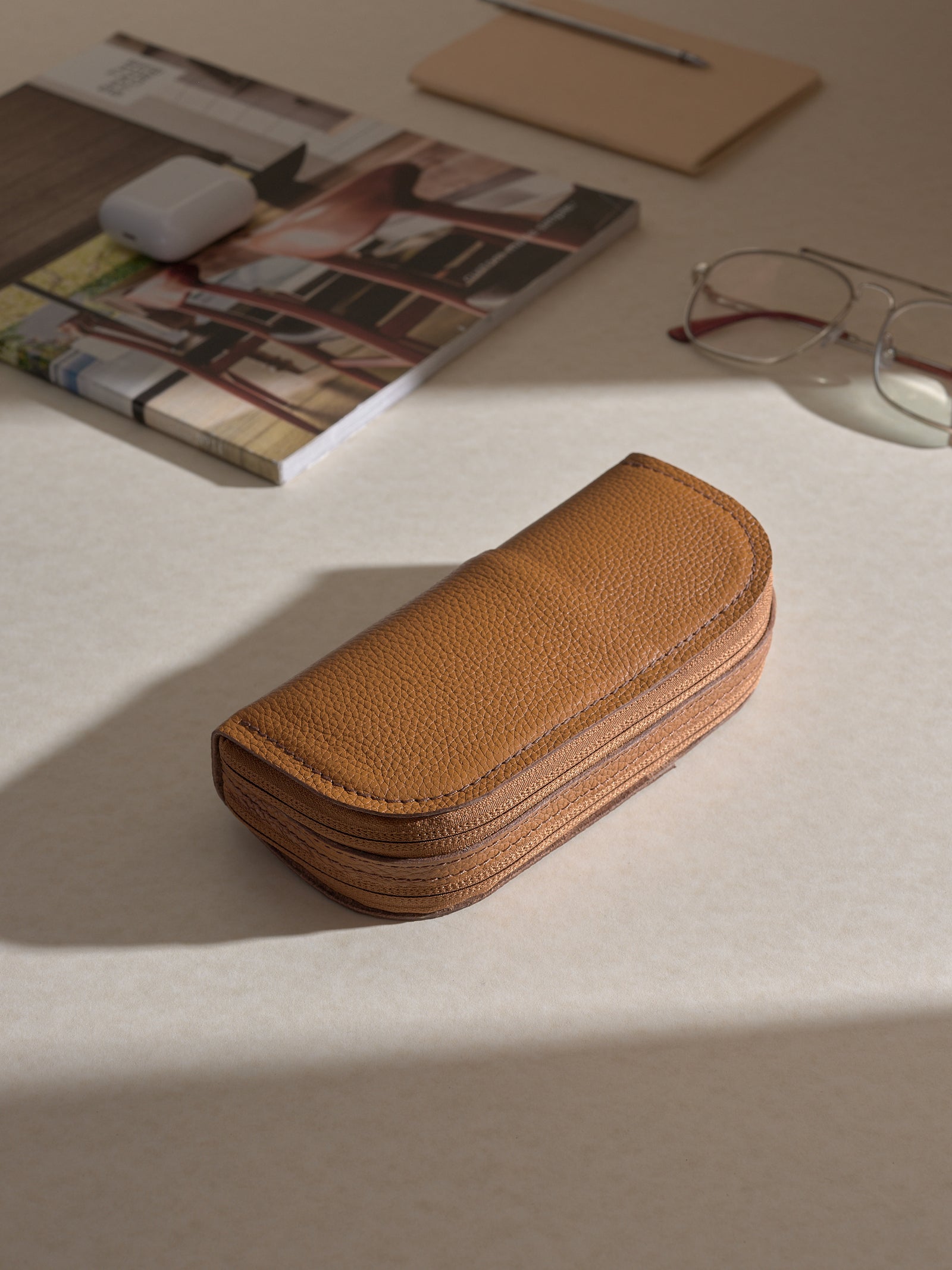 The tan leather double glasses case lying on a light-colored desk surface, surrounded by other items like a magazine, a notepad, a pen, and a pair of glasses.