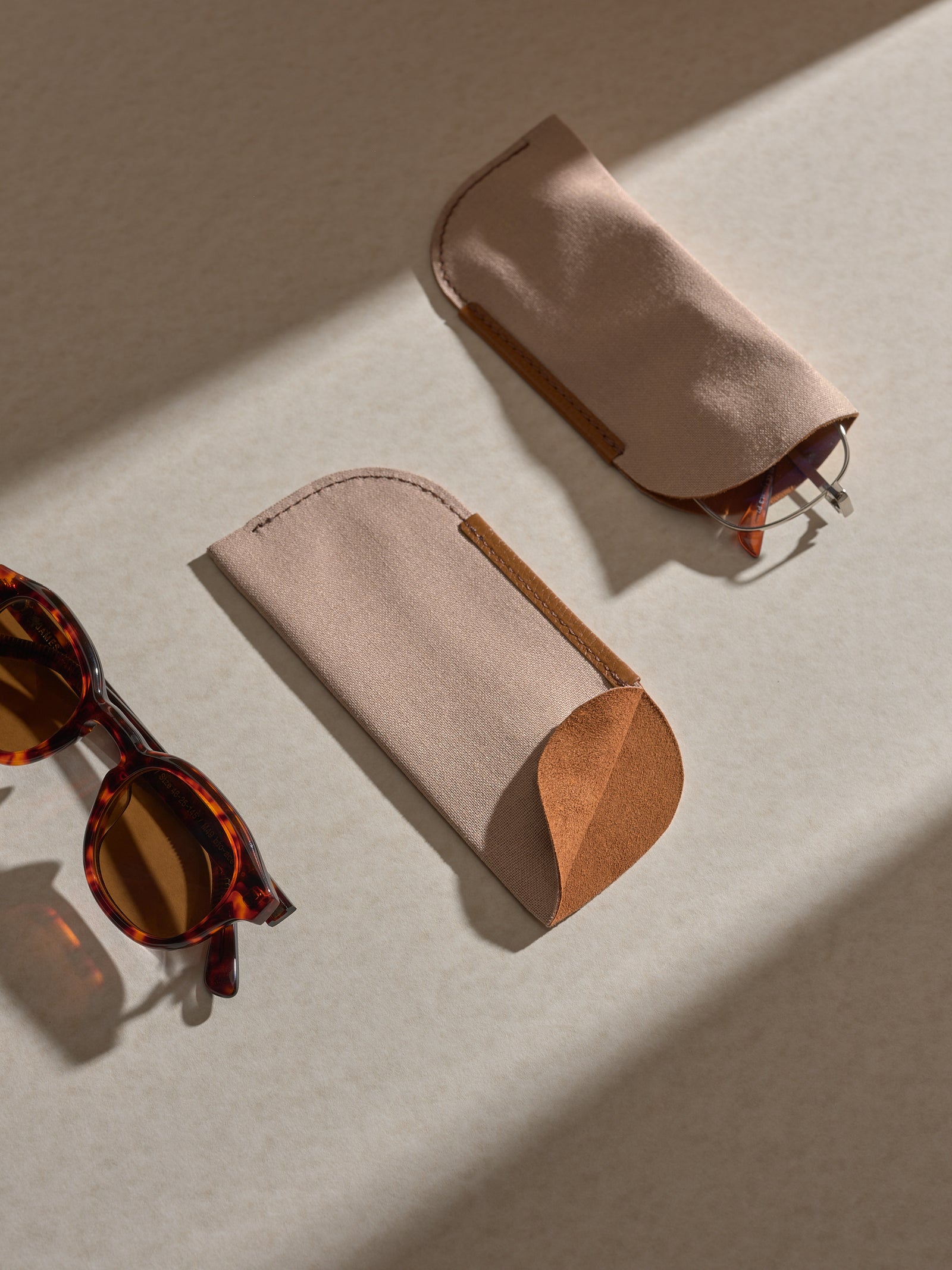 Two separate soft fabric sleeves, which hold the glasses within the main case, are shown on a light surface, with a pair of tortoise-shell sunglasses next to them.