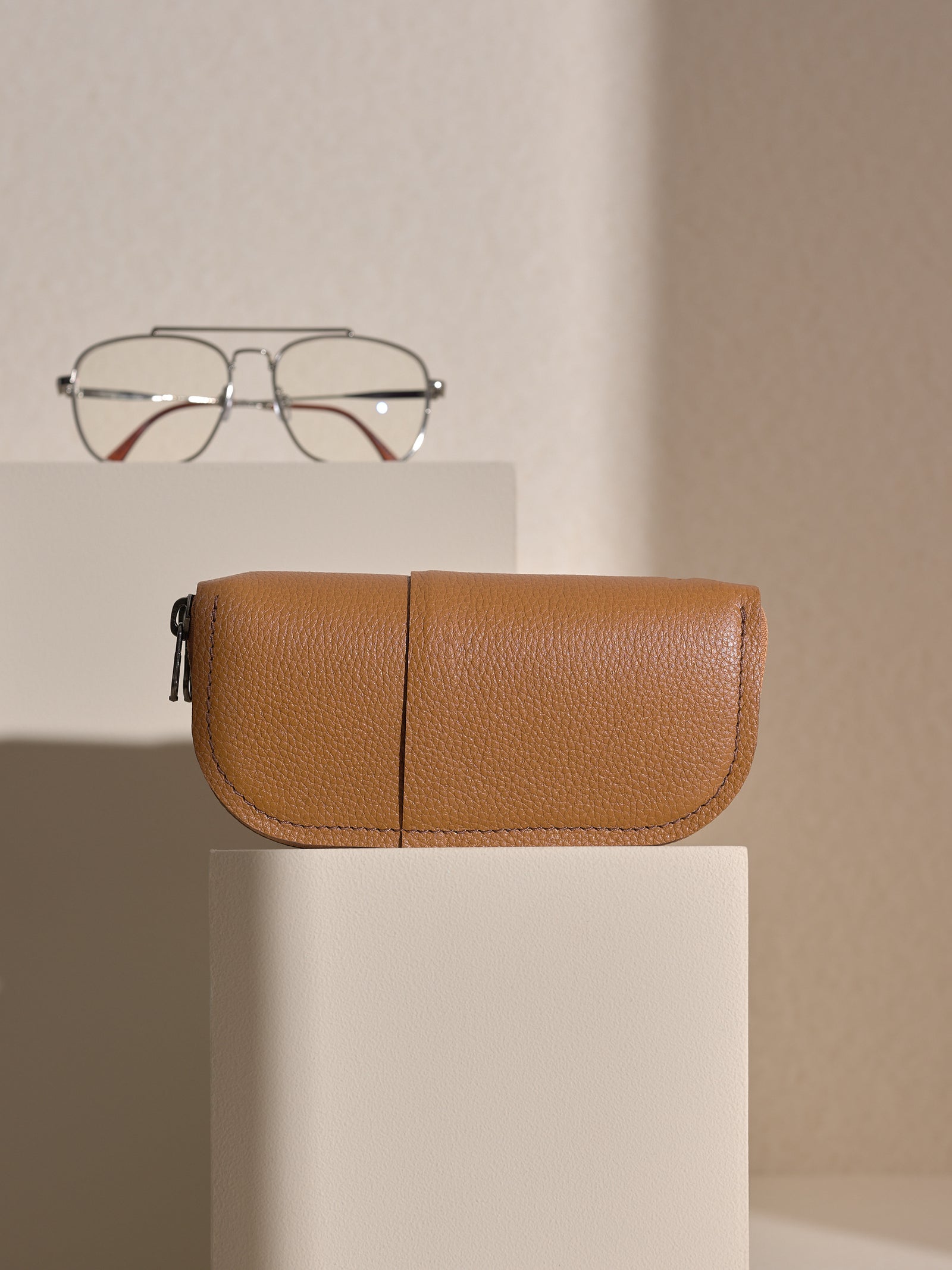 The tan Capra leather double glasses case resting horizontally on two white platforms, with a pair of silver-framed glasses positioned behind it on the upper platform.
