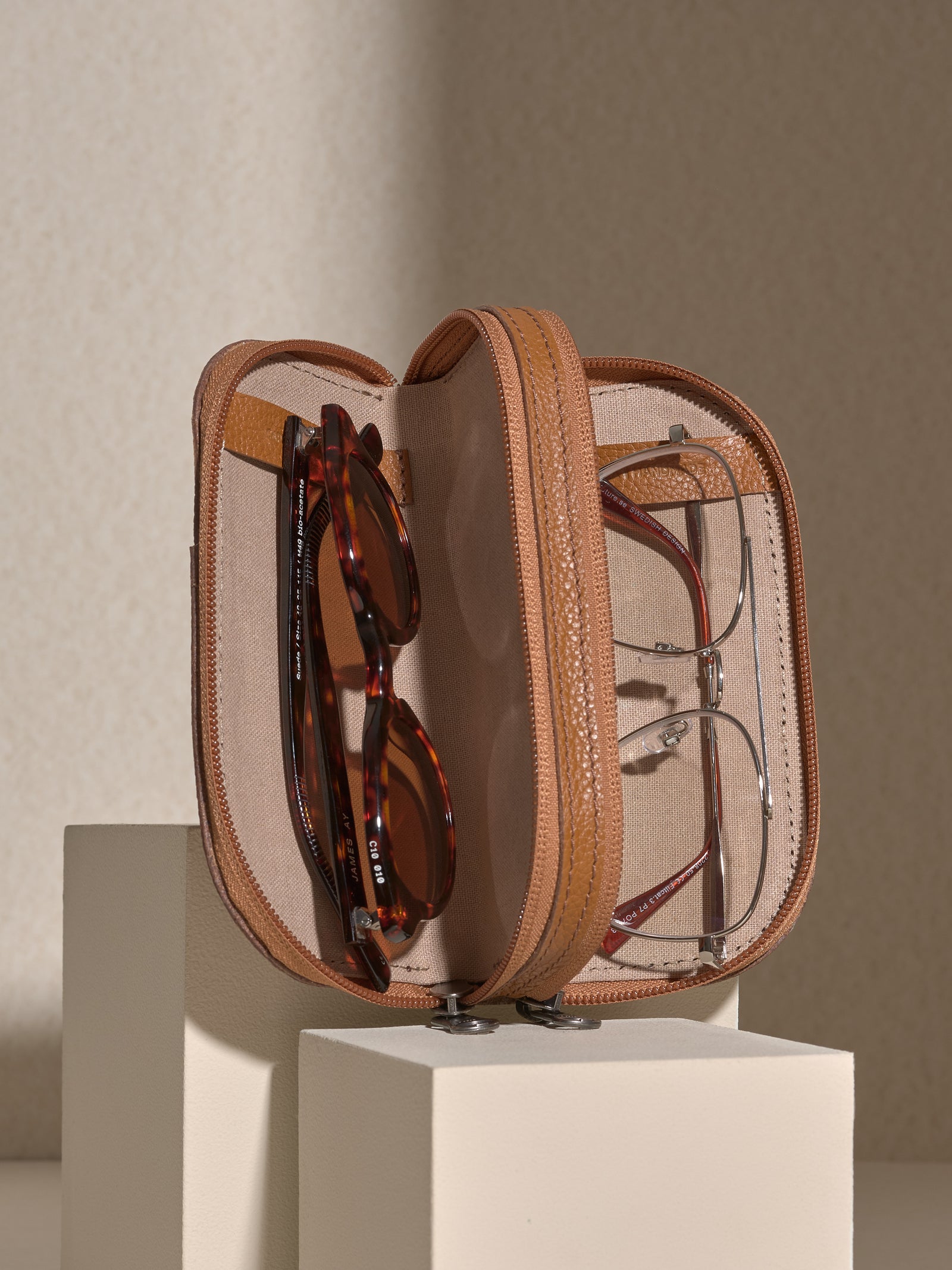 The zipped tan leather double glasses case opened to show two pairs of glasses stored inside: tortoise-shell sunglasses in one compartment and silver-framed clear glasses in the other.