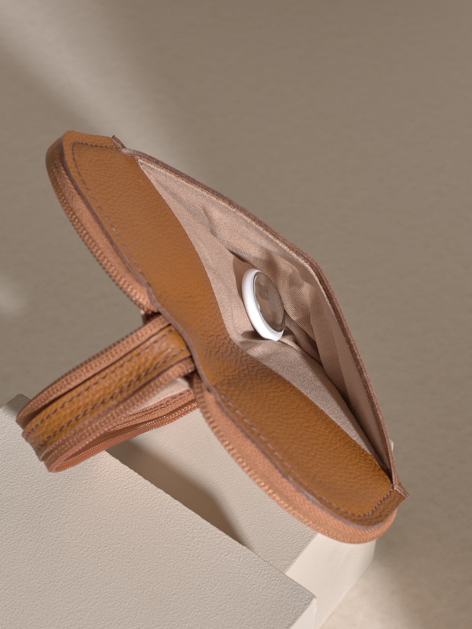 A detail shot of the opened, zippered tan leather glasses case, showing a small white circular object (likely an AirTag or tracker) placed inside a slip pocket in the lining.