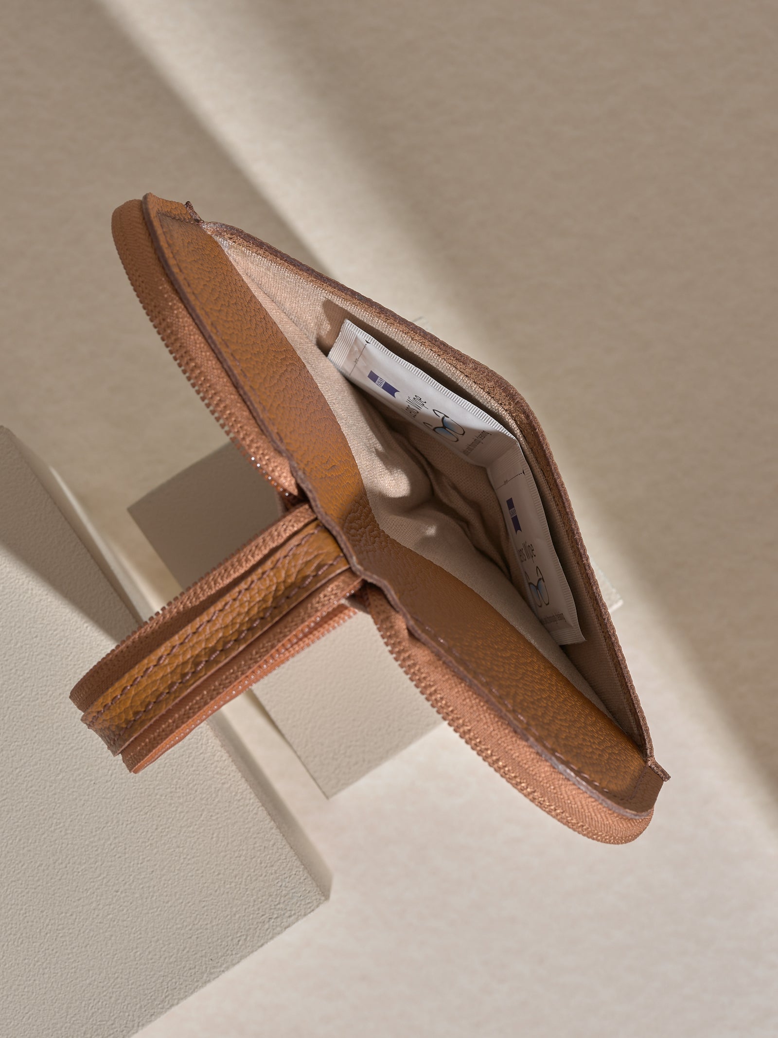 A detail shot of the opened, zippered tan leather glasses case, showing a small white circular object (likely an AirTag or tracker) placed inside a slip pocket in the lining.