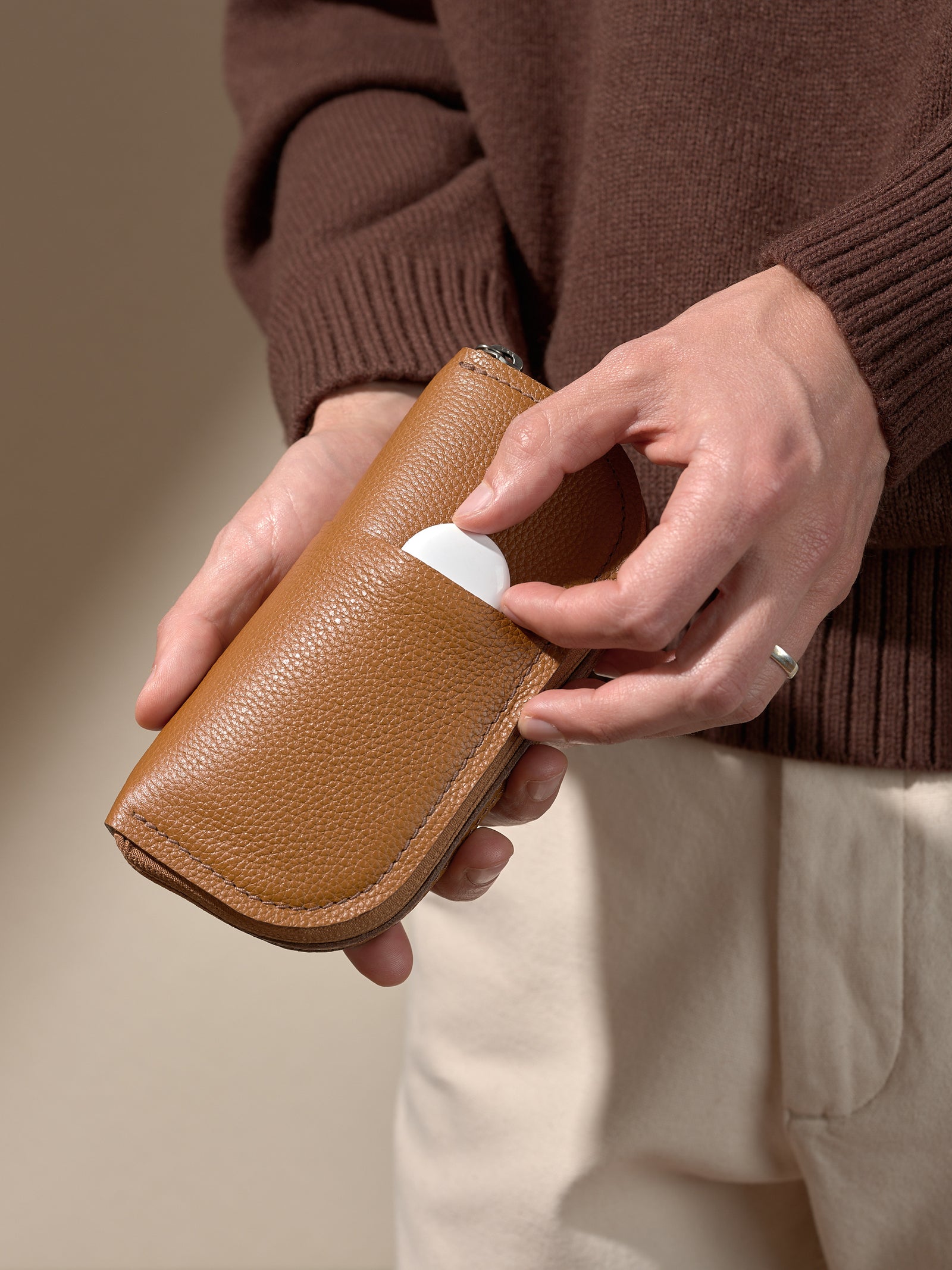 A person's hands holding the closed, textured tan leather glasses case and placing a small, white, circular object (possibly an AirTag or earbud case) into a small exterior slip pocket
