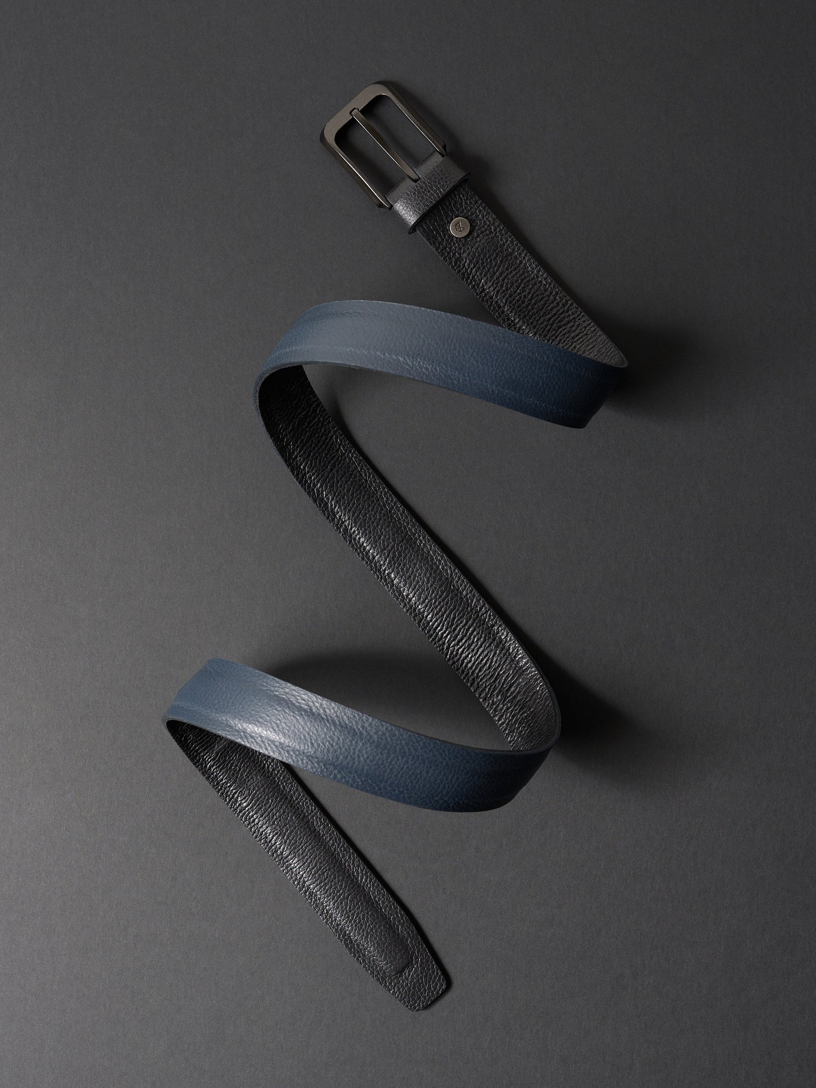 Overhead view of a coiled double-sided 32mm leather belt in black and navy on a dark gray background.