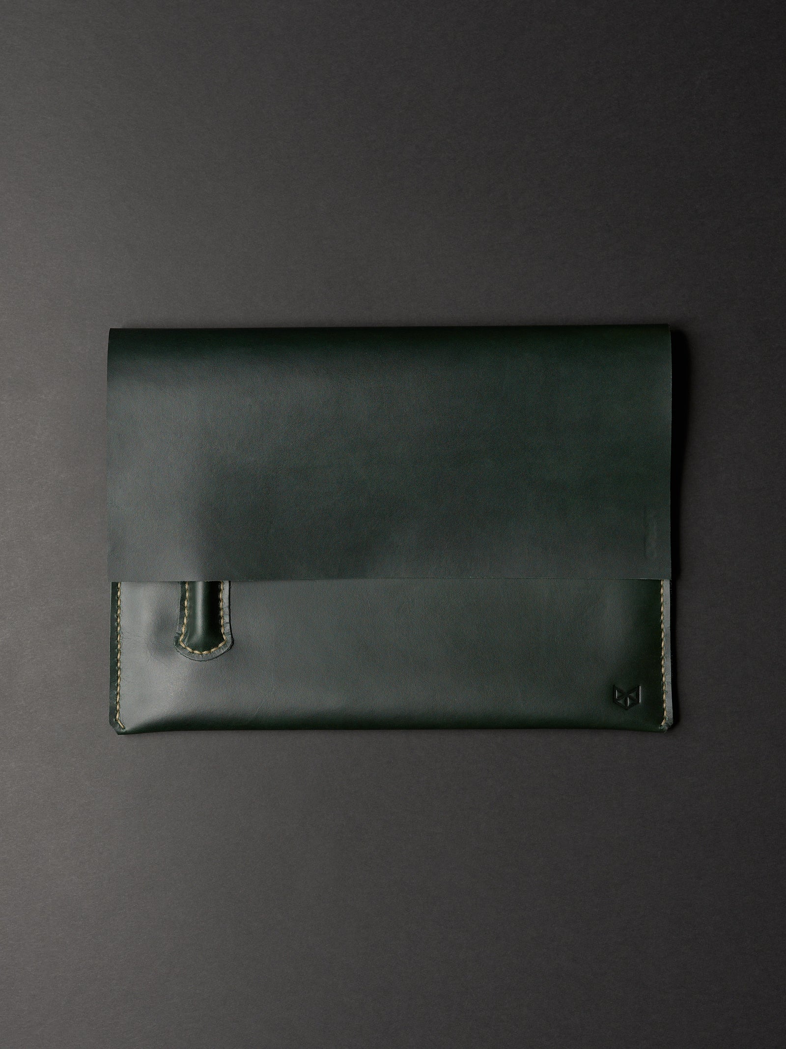 An overhead view of a closed, forest green leather laptop sleeve with a subtle logo in the corner on a dark background.