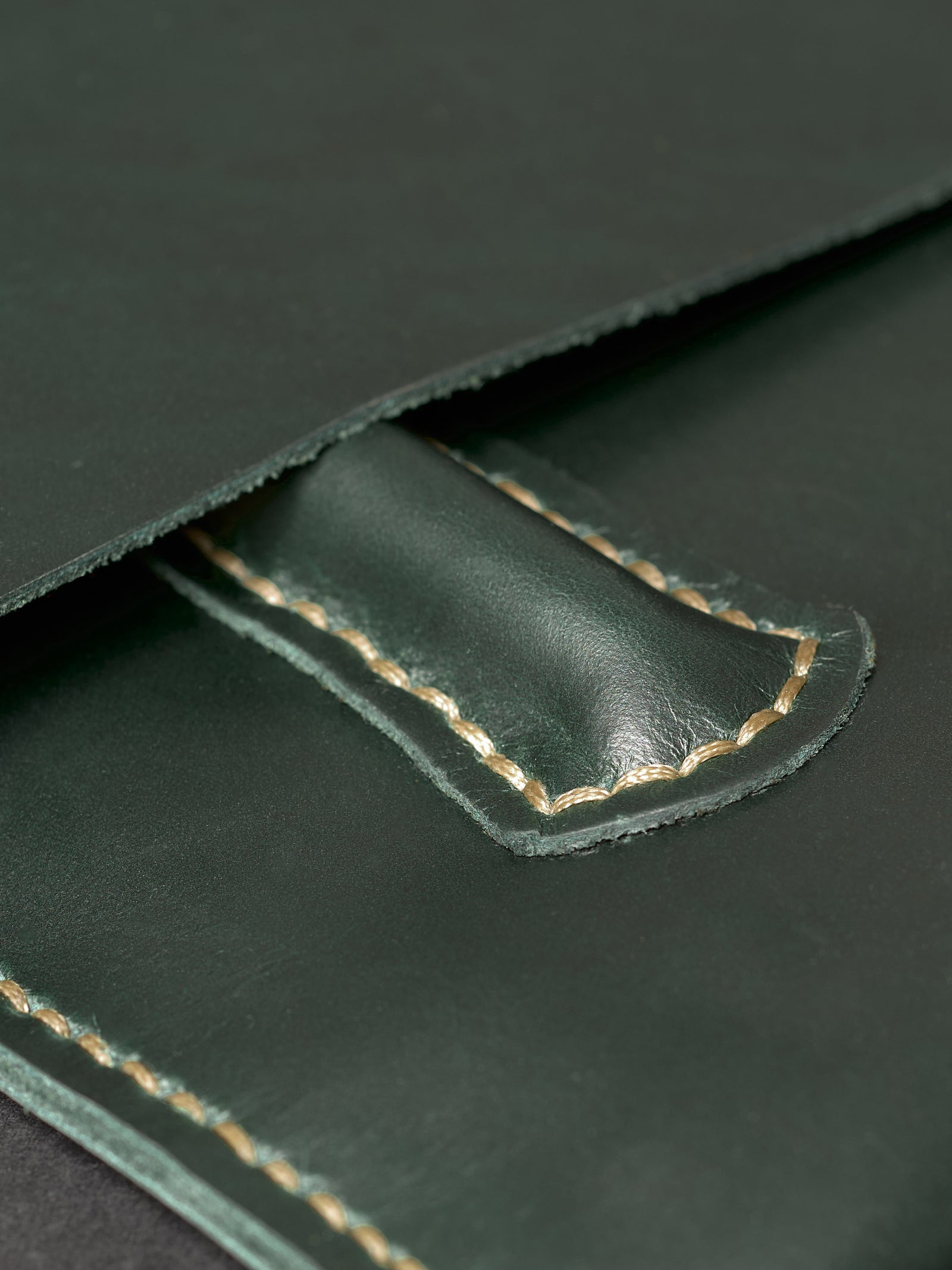 Extreme close-up of a forest green leather pen loop with intricate, hand-stitched tan thread on a dark surface.