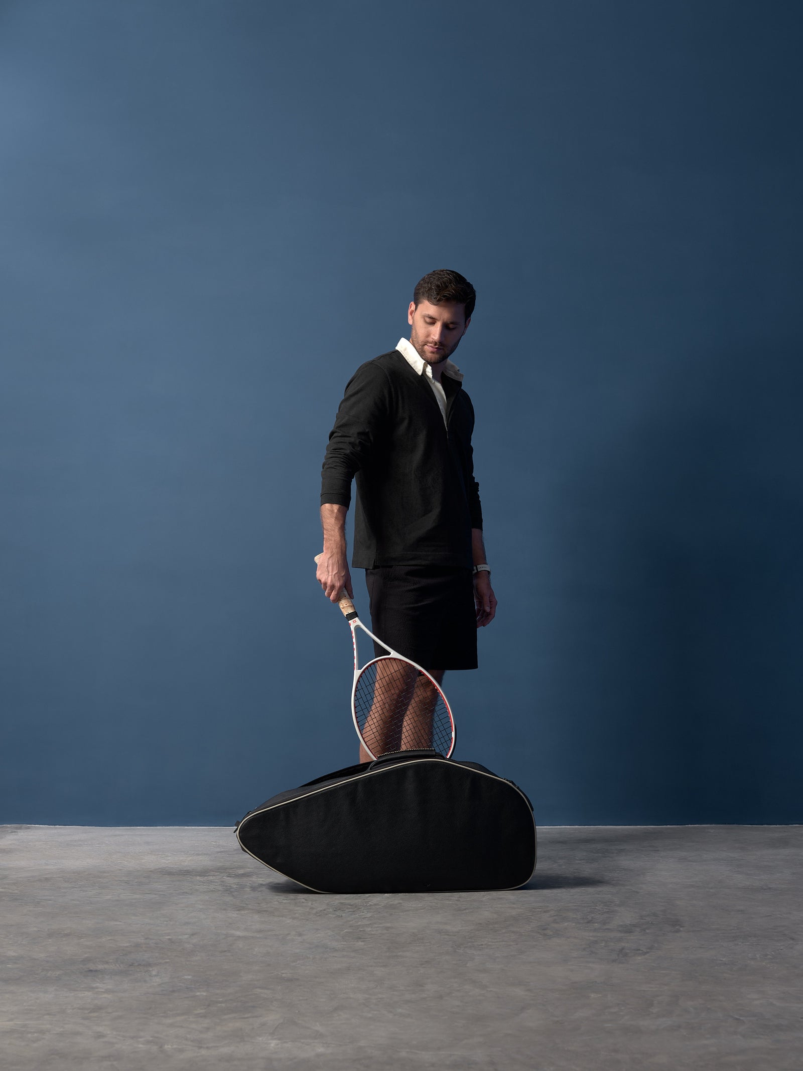 Full-length shot of a man standing with a white tennis racquet and a black leather thermobag.