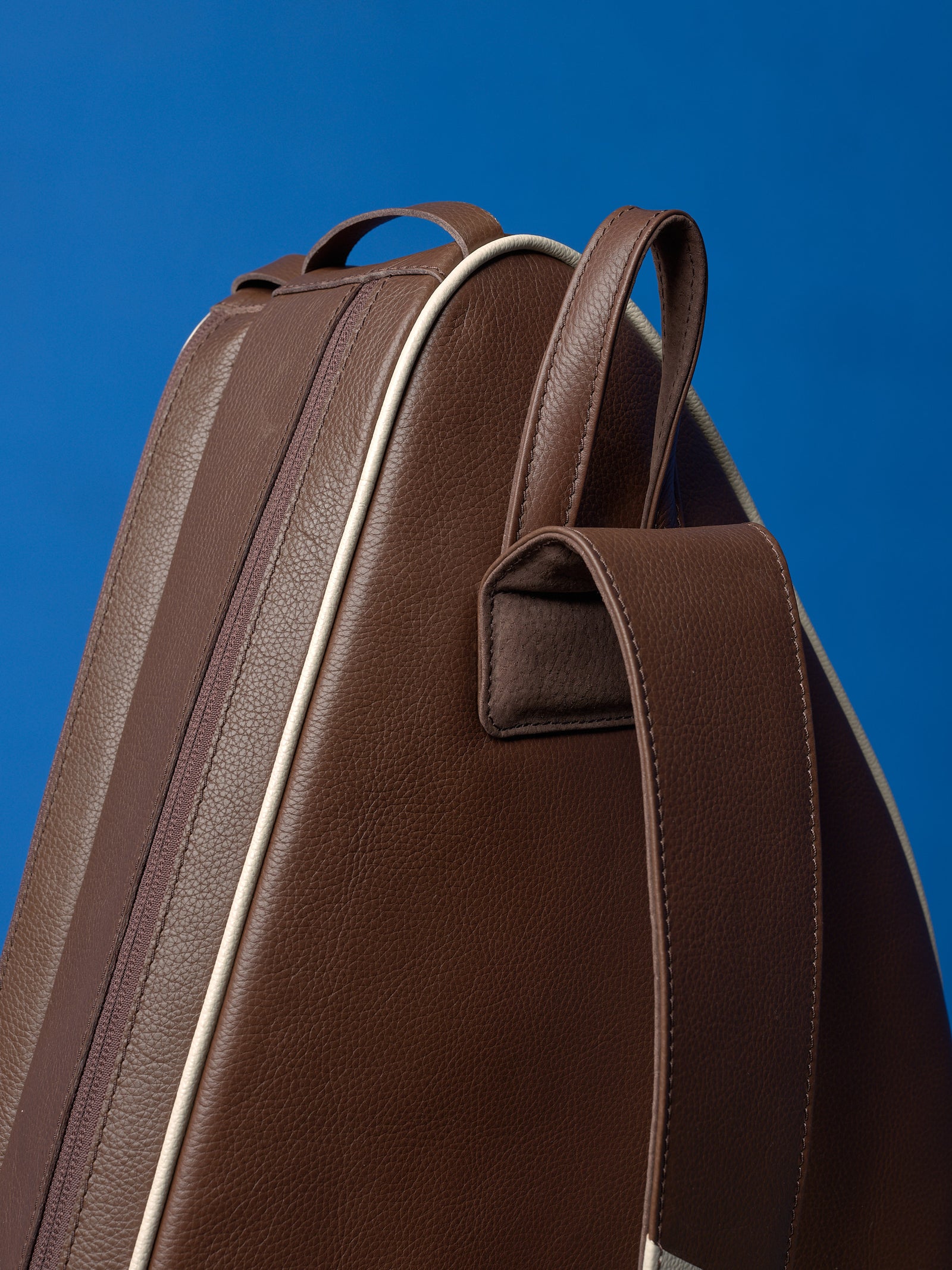 Close-up of the top handle and reinforced stitching on a handcrafted leather tennis bag against a blue background.