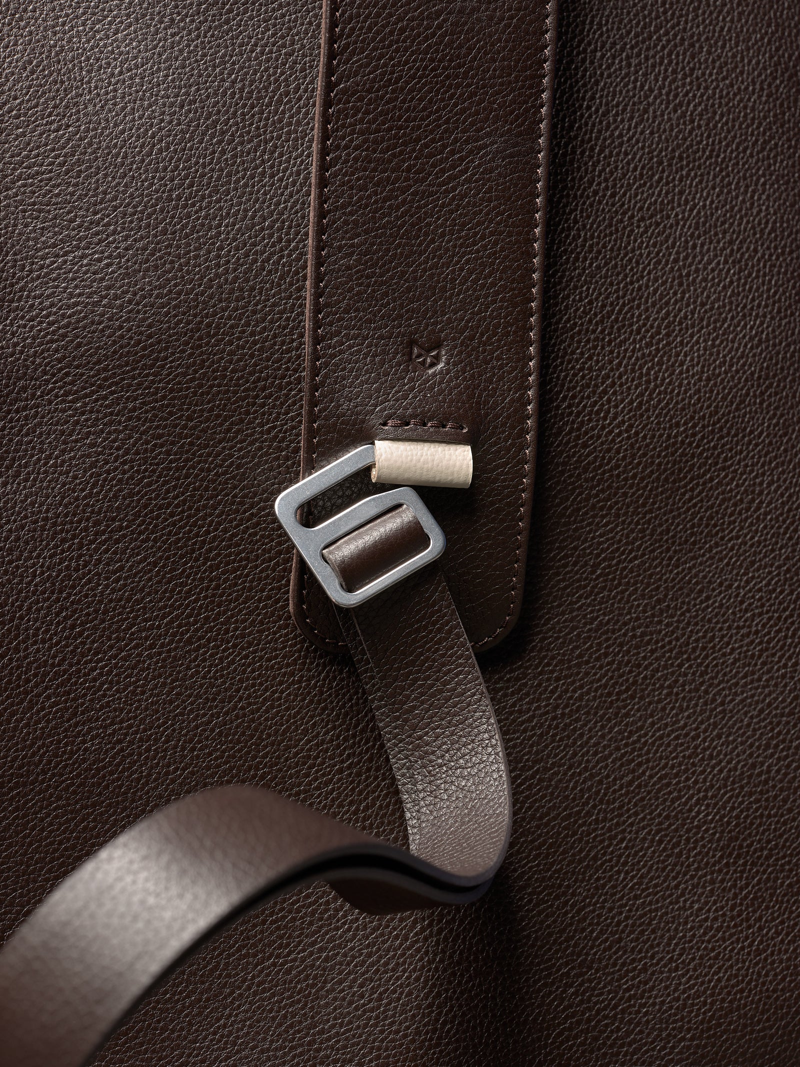 Close-up of a dark brown pebbled leather bag, featuring a metallic buckle, a leather strap, and a subtle stamped logo.