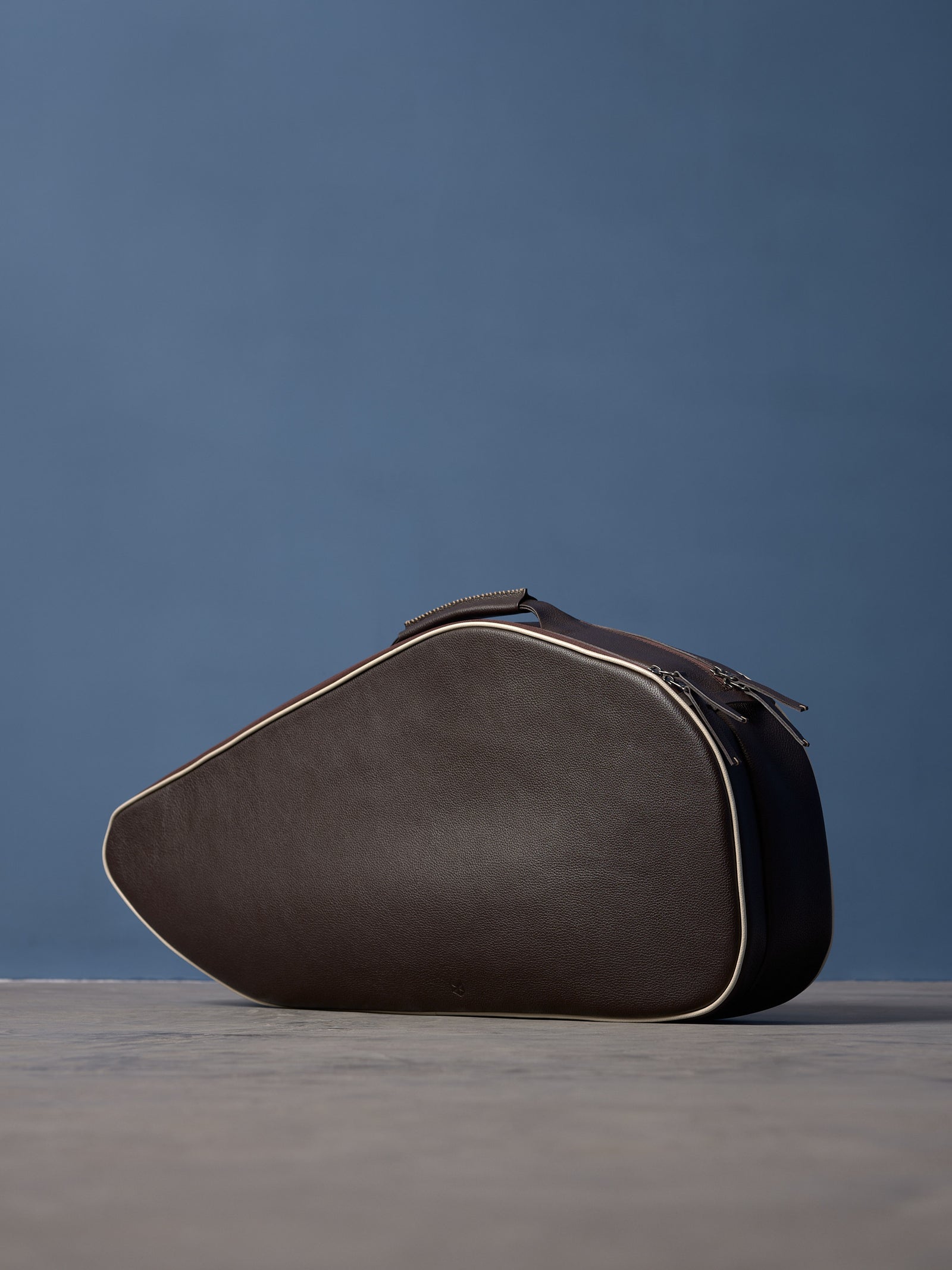 Premium brown leather racket bag with cream trim and dual zips, presented on a minimalist grey surface against a blue backdrop.
