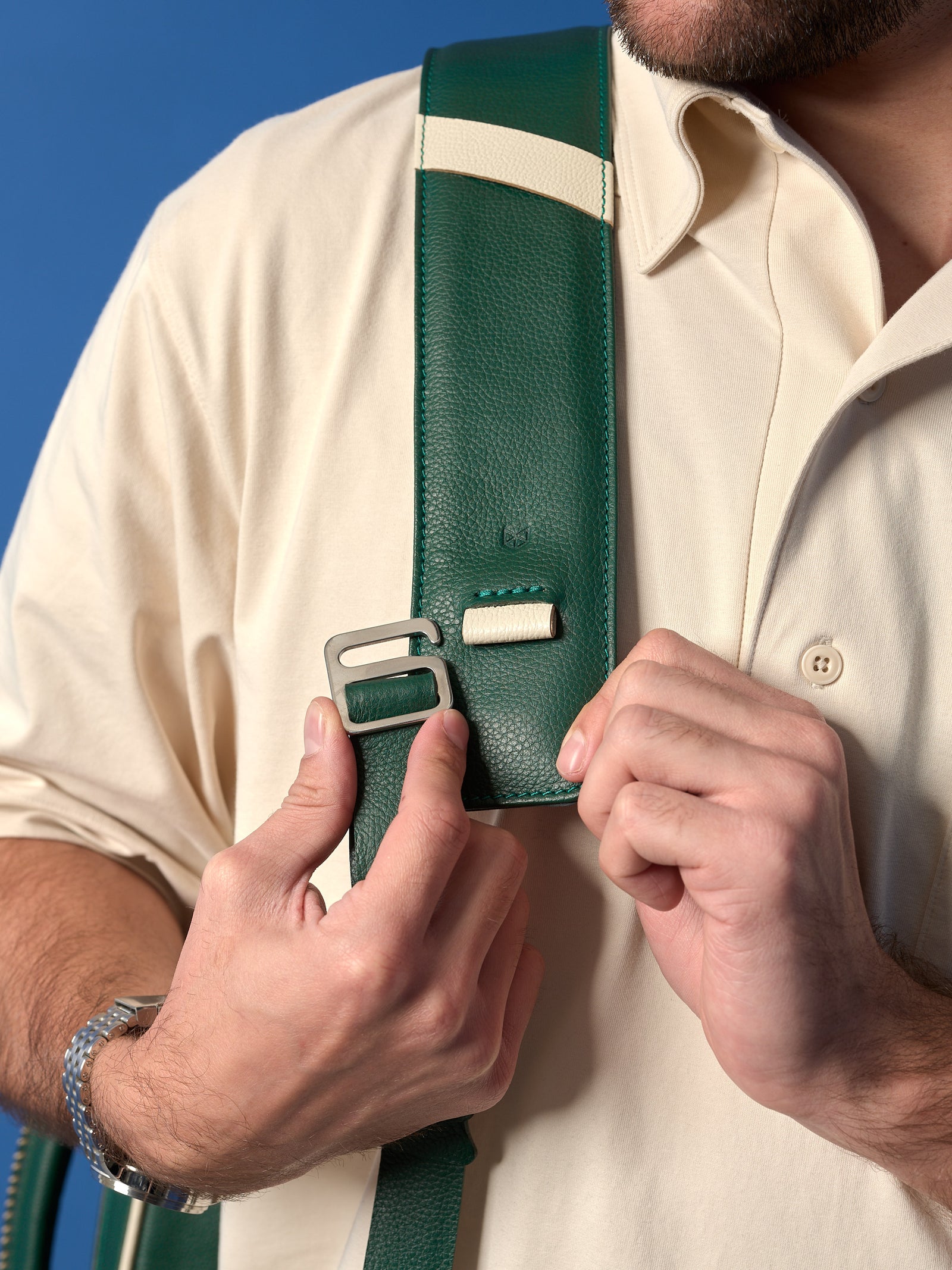 A close-up of a man’s hands adjusting a silver metal G-hook buckle on the dark green leather shoulder strap, showing a contrast cream leather accent and pebble-grain texture.