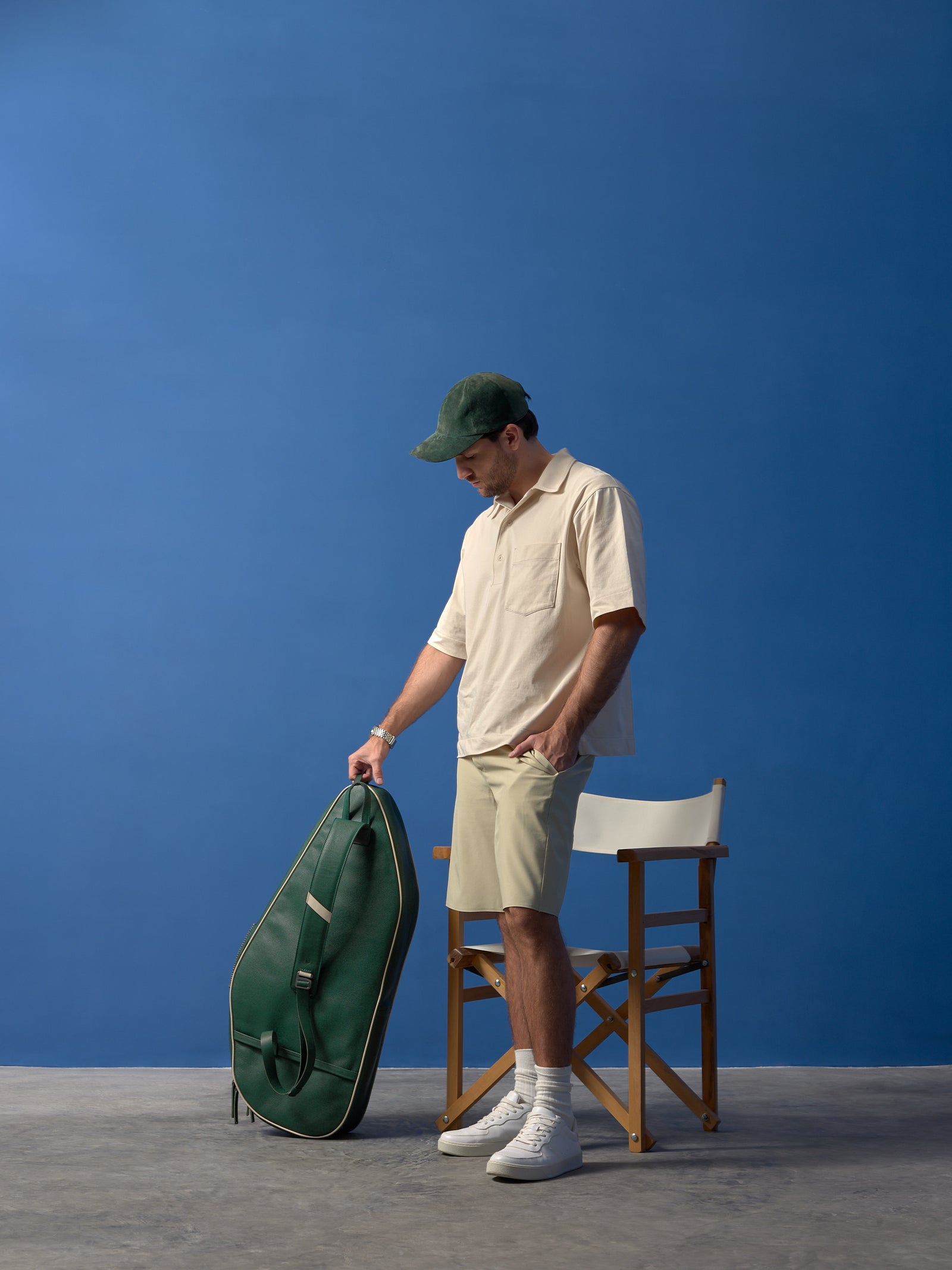 A full-body lifestyle shot of a man standing next to a classic wooden director’s chair, holding the dark green leather tennis bag against a cobalt blue studio wall.