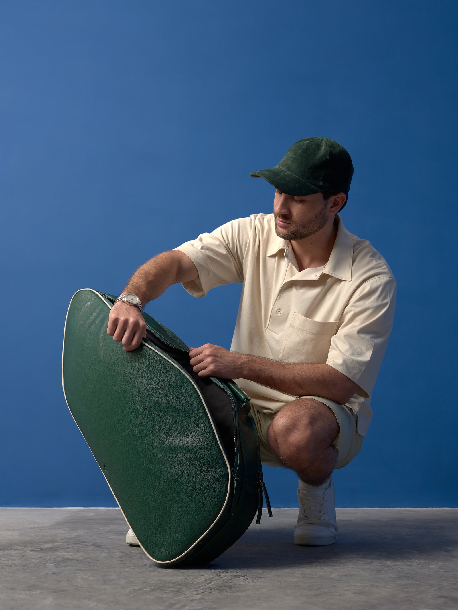 A man in a beige knit polo and a green suede cap crouches, adjusting the metal zipper of a dark green leather tennis bag with white piping and leather zipper pulls.