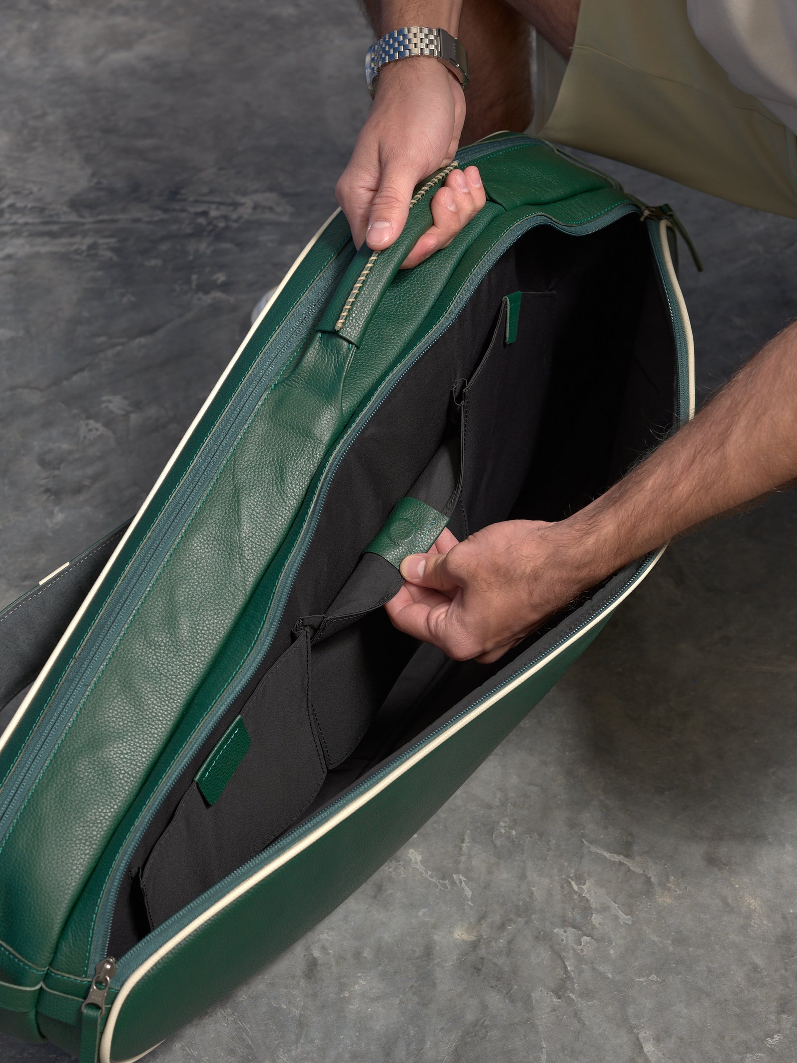 An over-the-shoulder view of a hand opening an internal black pocket inside the dark green leather tennis bag. The white piping and sturdy leather construction are visible.