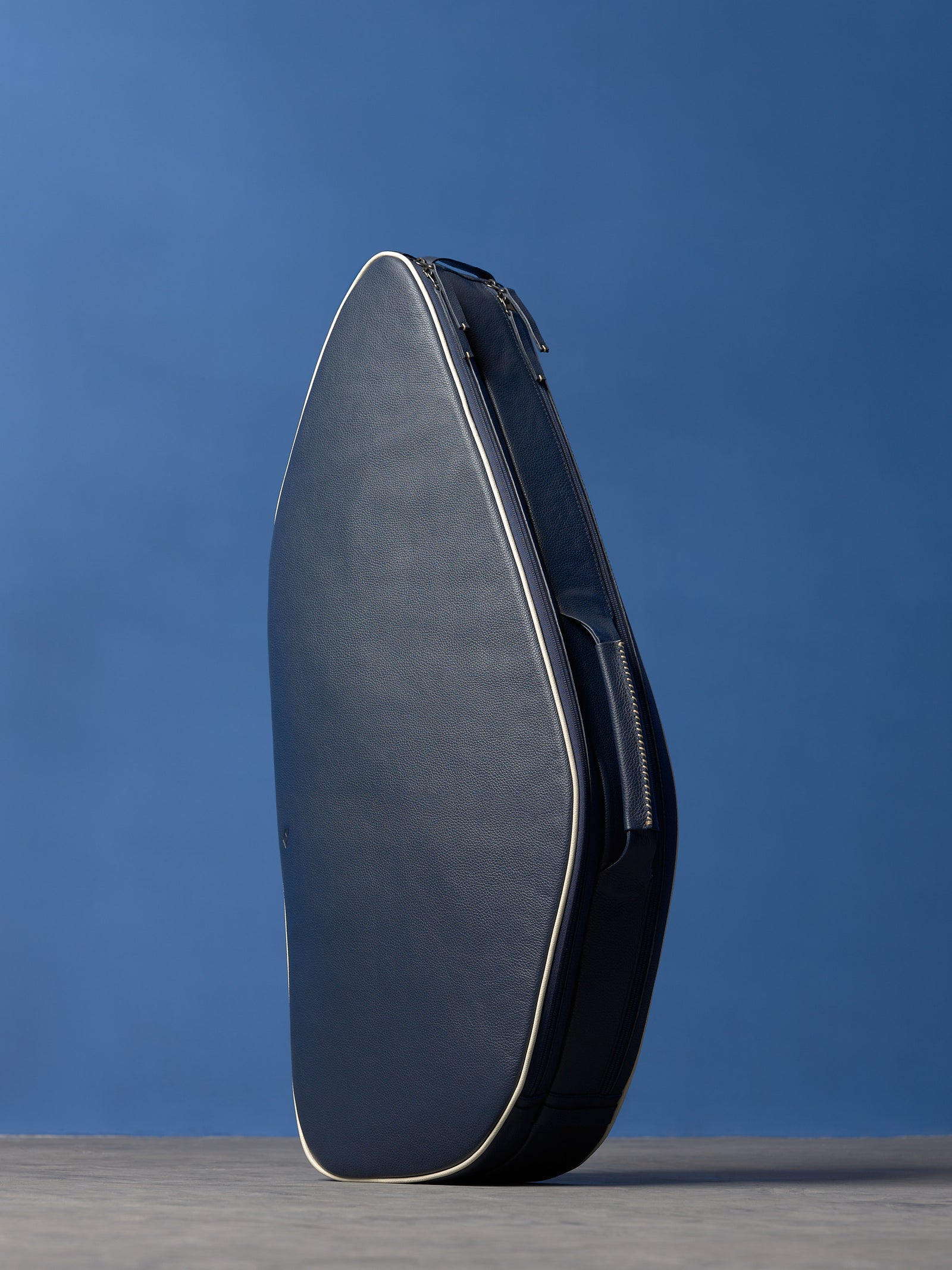 The navy blue leather tennis bag standing upright, showcasing its shape and structure against the blue background.
